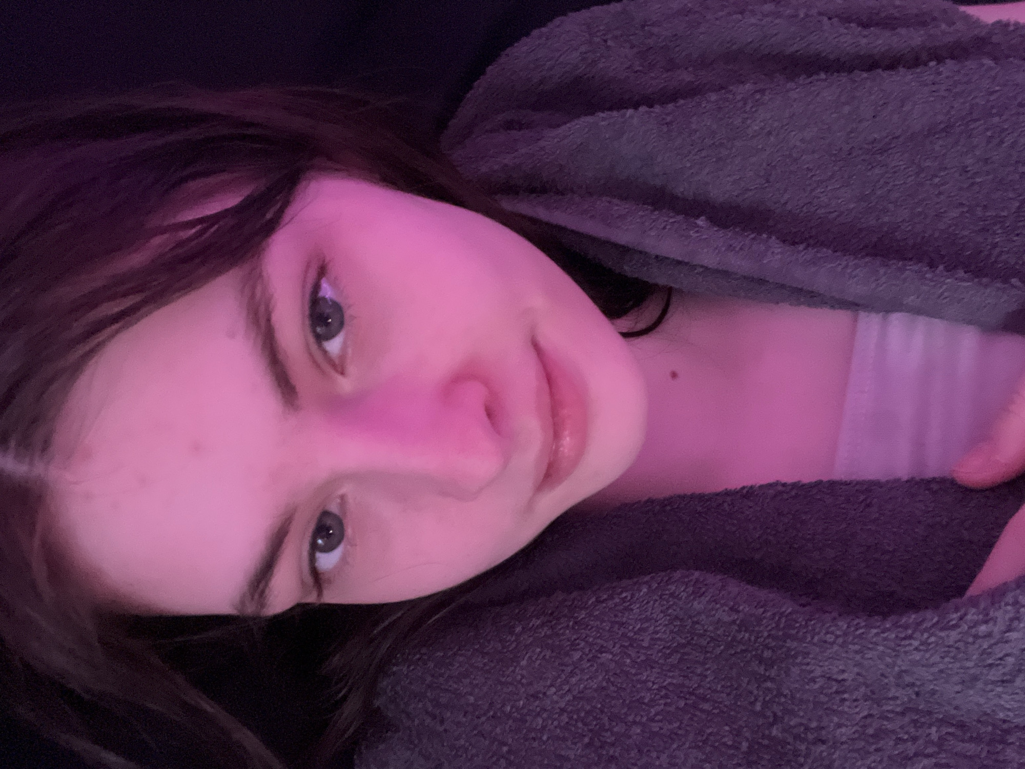 A young woman lies down, facing the camera with a slight smile. She has dark brown hair and fair skin, with a mole visible on her neck. She is wearing what appears to be a grey terrycloth robe or jacket over a light-colored garment. The lighting in the scene is pink or purple-toned. The image provides no information to place the location in Saint Petersburg, Russia, or any other specific location.