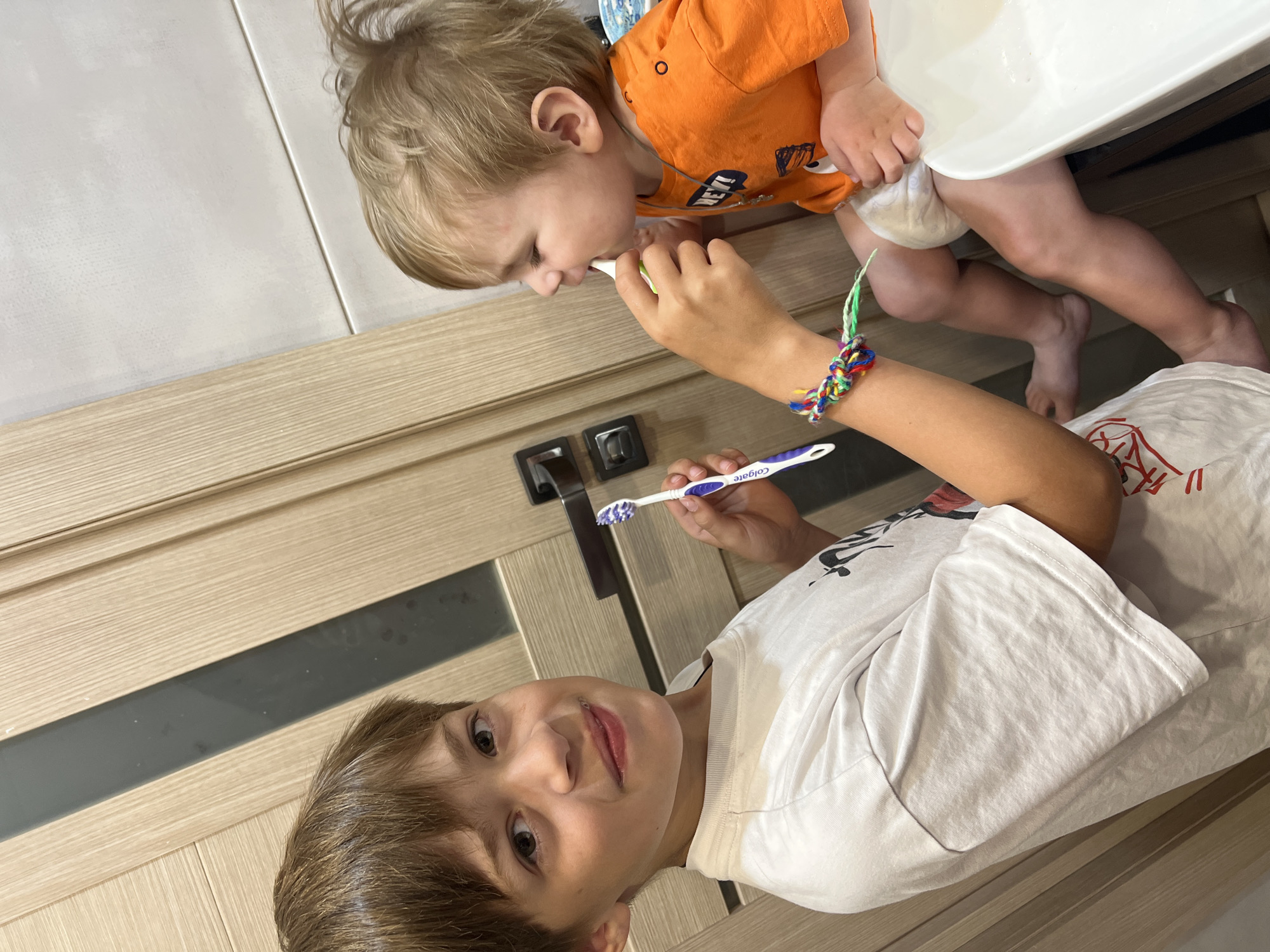 In this candid shot, two young boys are captured in a bathroom, likely during their morning or evening routine. The older boy, in the foreground, is wearing a white t-shirt and looking directly at the camera with a playful smile. He is holding a blue and white toothbrush. Beside him, the younger boy, dressed in an orange t-shirt and a diaper, is actively brushing his teeth. His head is tilted, focusing on the task at hand.
The scene is set against a backdrop of modern wooden cabinetry and white tiled walls, suggesting a clean and contemporary bathroom environment. A white basin is visible behind the younger boy, likely part of a sink or bathtub. The lighting is bright and natural, indicative of daytime. The overall mood is one of familial intimacy and everyday moments.
A notable detail is a colorful woven bracelet on the older boy's wrist, adding a touch of childlike personality to the scene. The visible text on the toothbrush reads "Colgate". The photograph, taken from a slightly elevated angle, emphasizes the interaction between the two brothers as they engage in the common activity of oral hygiene.