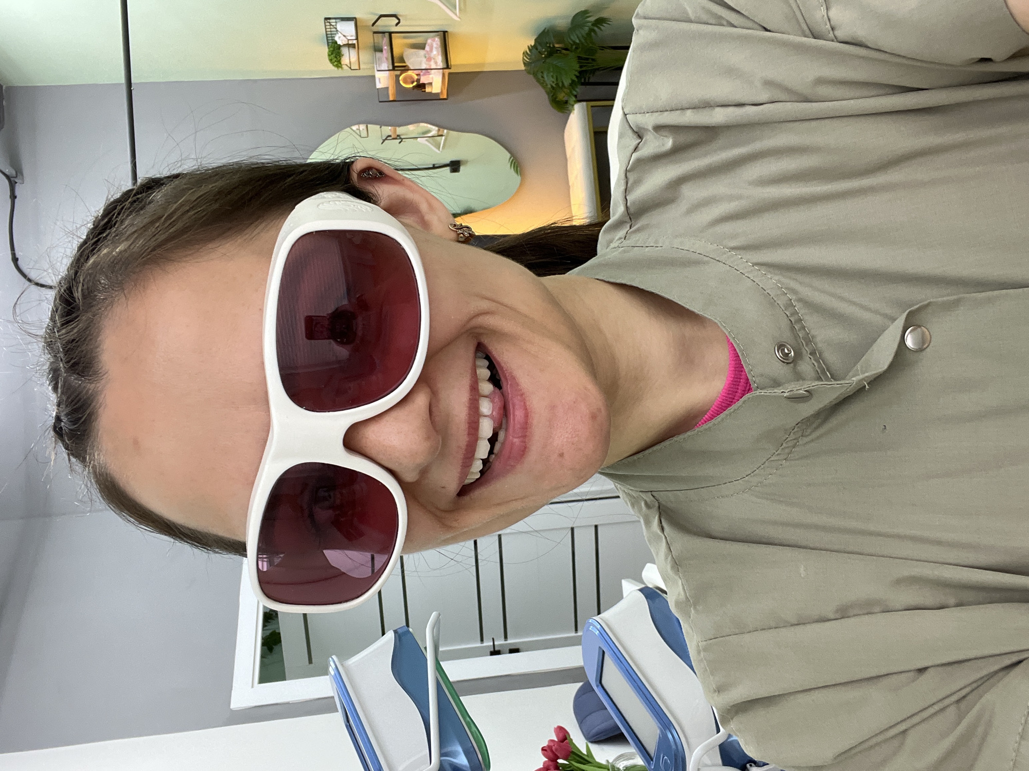 A woman with medium-length brown hair styled in a braid along her hairline is smiling broadly while wearing large, white sunglasses with burgundy lenses. She is wearing a light brown collared shirt or jacket with snap closures. The background appears to be an indoor setting. Visible elements include a white countertop with objects such as devices with blue accents, possibly part of dental or medical equipment. There is a small bouquet of red tulips on the counter. Behind her, a white-framed window is partially visible, and further back, there is a wall with a decorative mirror and shelving units. The image is rotated slightly, giving a tilted perspective of the scene. The location is Chaikovsky, Russia.