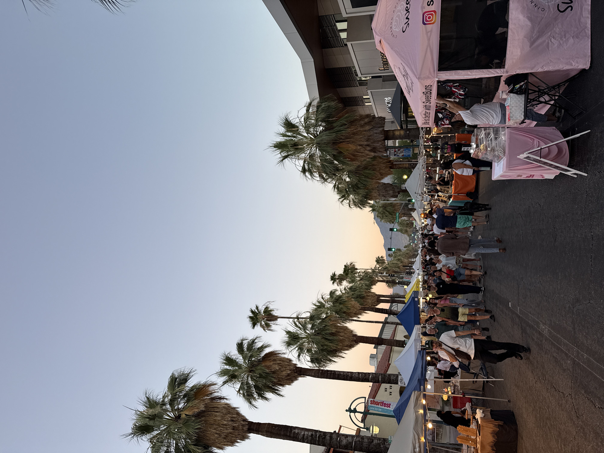 The image depicts a street fair or market scene in Palm Springs, California, United States of America, at dusk. A street lined with palm trees and storefronts is filled with people browsing market stalls. On the right side of the street, a pink tent with the branding "SweetBuns" is visible, displaying small items for sale. A woman is attending to the stall. On the left side, several other market tents are set up along the sidewalk. People are walking down the street, some individually and some in groups. The sky is a pale blue with hints of orange near the horizon, indicating the setting sun. Mountains are visible in the distance. The overall atmosphere suggests a lively outdoor event.