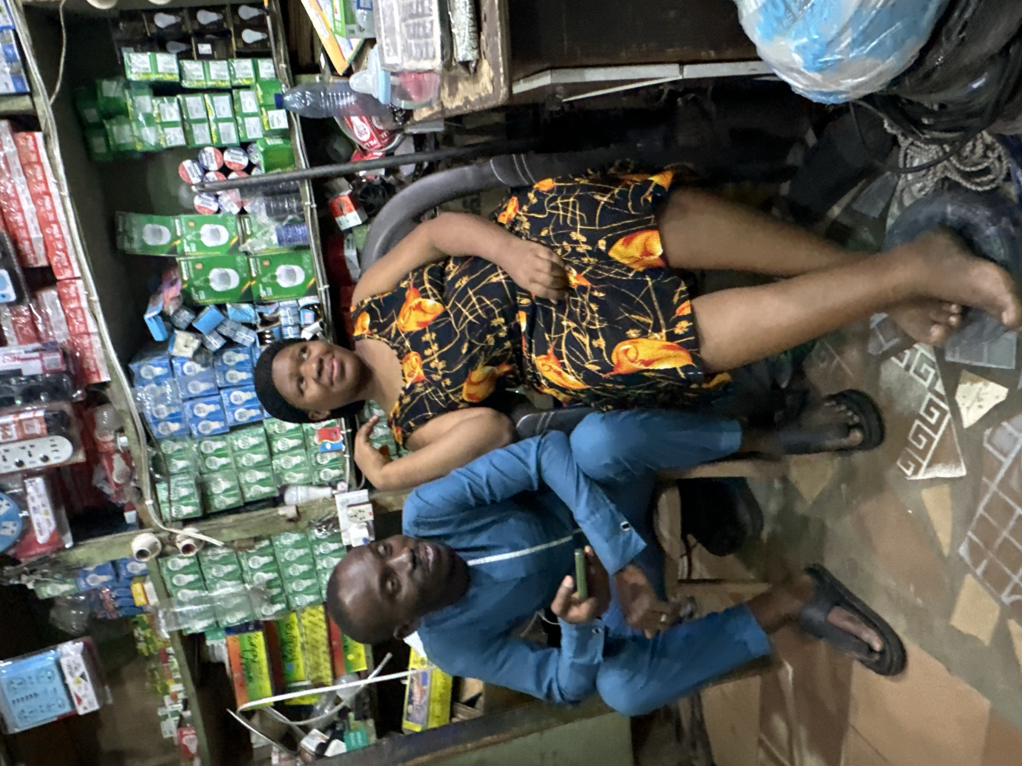 This image captures an indoor scene within a retail shop in Narayi, Nigeria. Two individuals, a man and a woman, are seated in the foreground, facing slightly towards each other.
The man, positioned on the left, has dark skin and short dark hair. He is wearing a long-sleeved, teal-blue traditional tunic with a vertical white stripe on the chest, and matching blue trousers. He wears black flip-flops. His left hand holds a dark-colored smartphone, and his right arm rests on his leg. He is looking towards the right with a slight smile.
The woman, seated on the right, also has dark skin and is wearing a black headwrap. She is dressed in a black sleeveless dress adorned with yellow and orange floral patterns. She is barefoot. Her right arm is bent, with her hand resting on her upper chest/shoulder, and her left hand rests on her lap. She is looking towards the left with a smile.
Behind the individuals, wooden shelves are densely packed with merchandise, primarily packaged electrical components. Visible items include numerous green and blue boxes, many depicting light bulbs, as well as rolls of electrical tape, remote controls, and various other packaged goods. A small, rustic wooden table is to the right of the woman, holding a clear plastic water bottle and a baby bottle. The floor is tiled with terracotta-colored squares and lighter tiles featuring a white geometric (Greek key) pattern. Miscellaneous wires, plastic bags, and discarded items are on the floor near the woman's feet. The lighting suggests an interior, possibly artificial, setting.