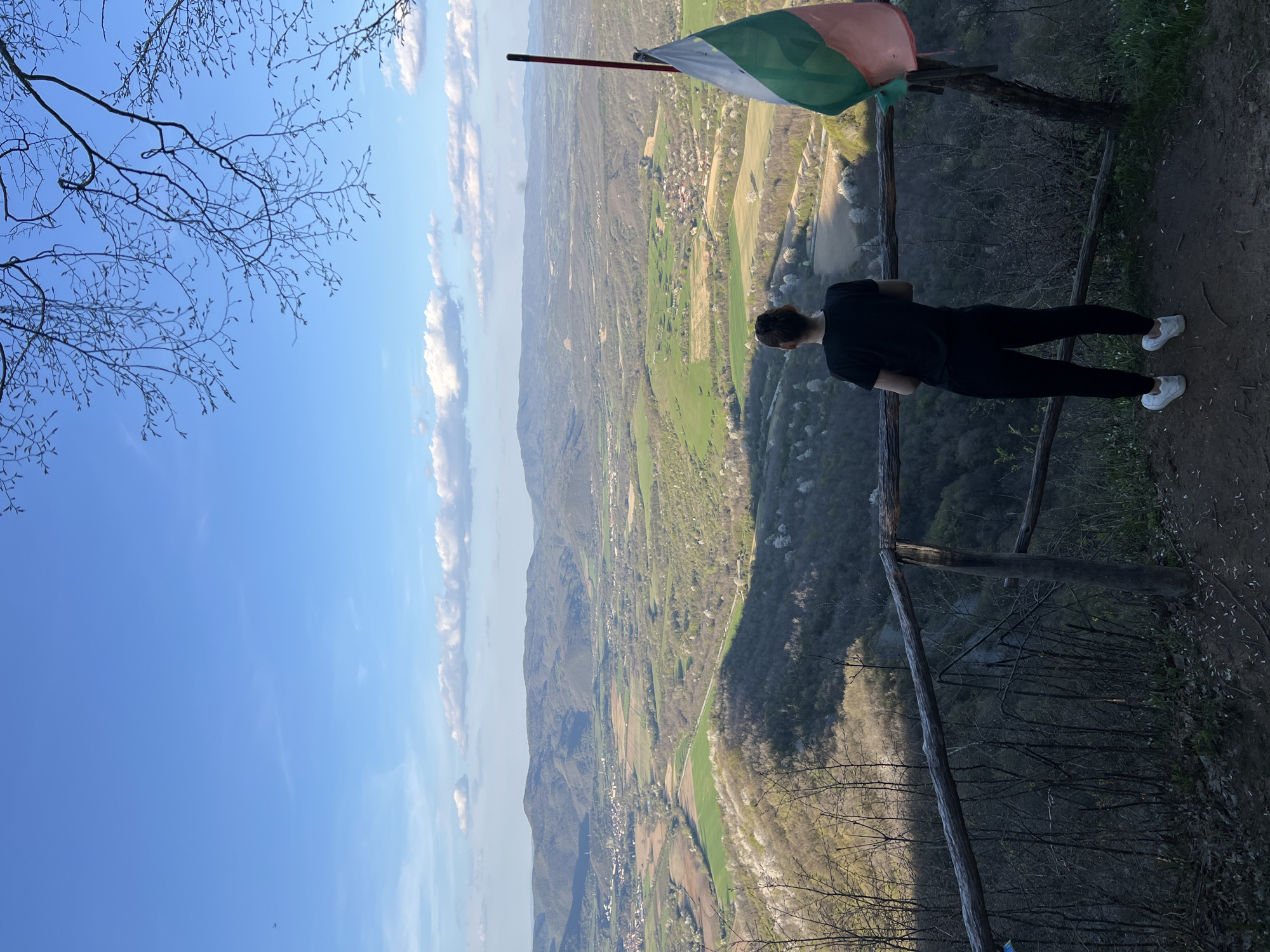 A landscape view, taken outdoors in daylight, depicts a woman standing on a hillside in Elena, Bulgaria. She is positioned near a wooden fence, gazing out at a valley, forests, and fields. The Bulgarian flag is mounted nearby on a pole. The sky is blue with scattered clouds. Tree branches are visible in the upper left corner.