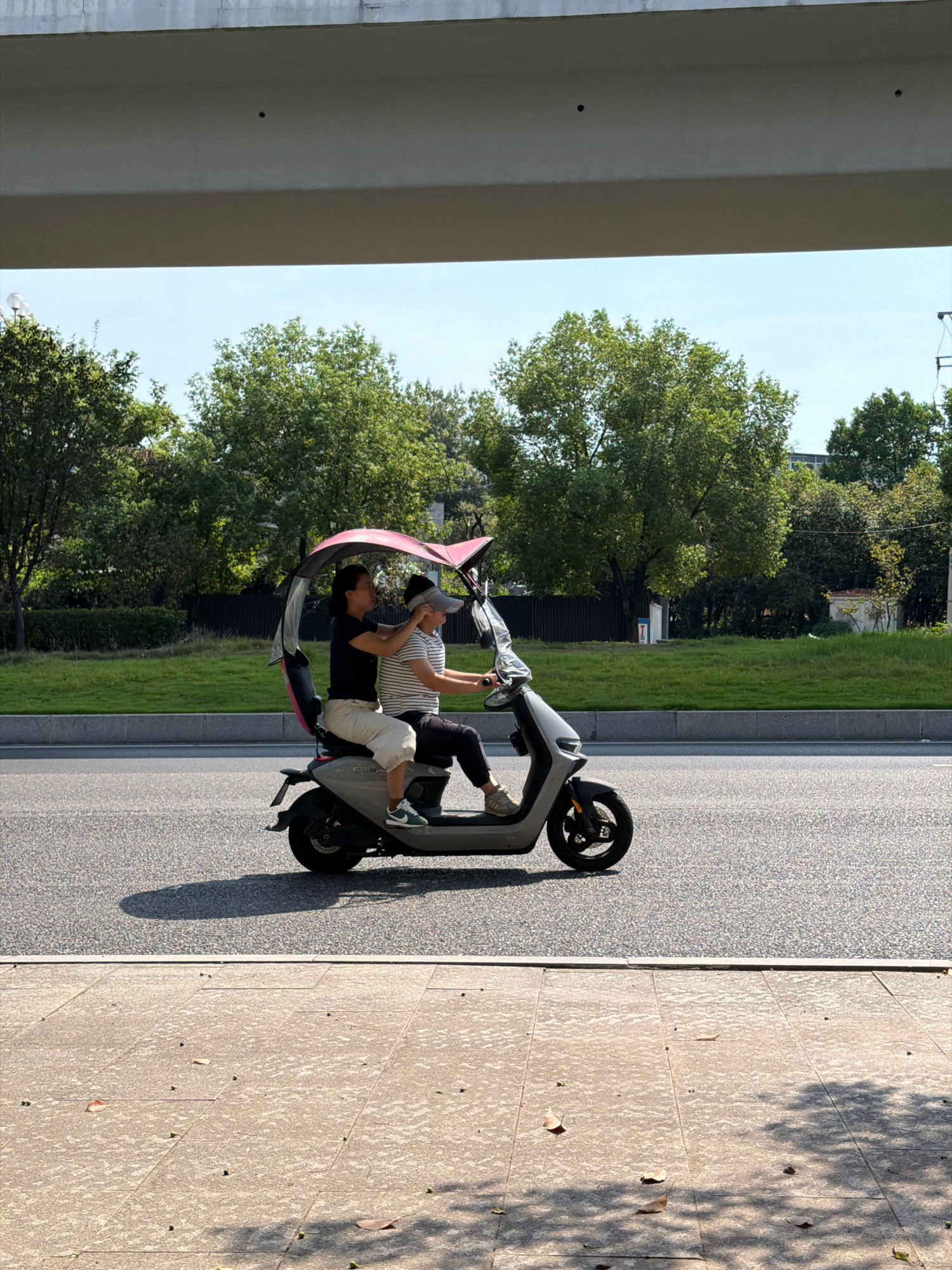 Two individuals are riding on an electric scooter under a pink canopy on a road in the city of Jiangjin, China. The person in front is wearing a black t-shirt and light-colored pants, while the person in the back is wearing a striped t-shirt and dark pants and a grey visor. They are both looking forward. The scooter is grey and has a single wheel visible at the front and rear. The road is paved with asphalt. To the left of the road is a sidewalk made of textured concrete with scattered leaves. To the right of the road, there is a grassy area bordered by a dark fence and then trees. In the background, there are more trees and some buildings. An elevated structure, possibly a bridge or overpass, is visible at the top of the image, casting a shadow on the road and sidewalk. The sky is clear and blue.