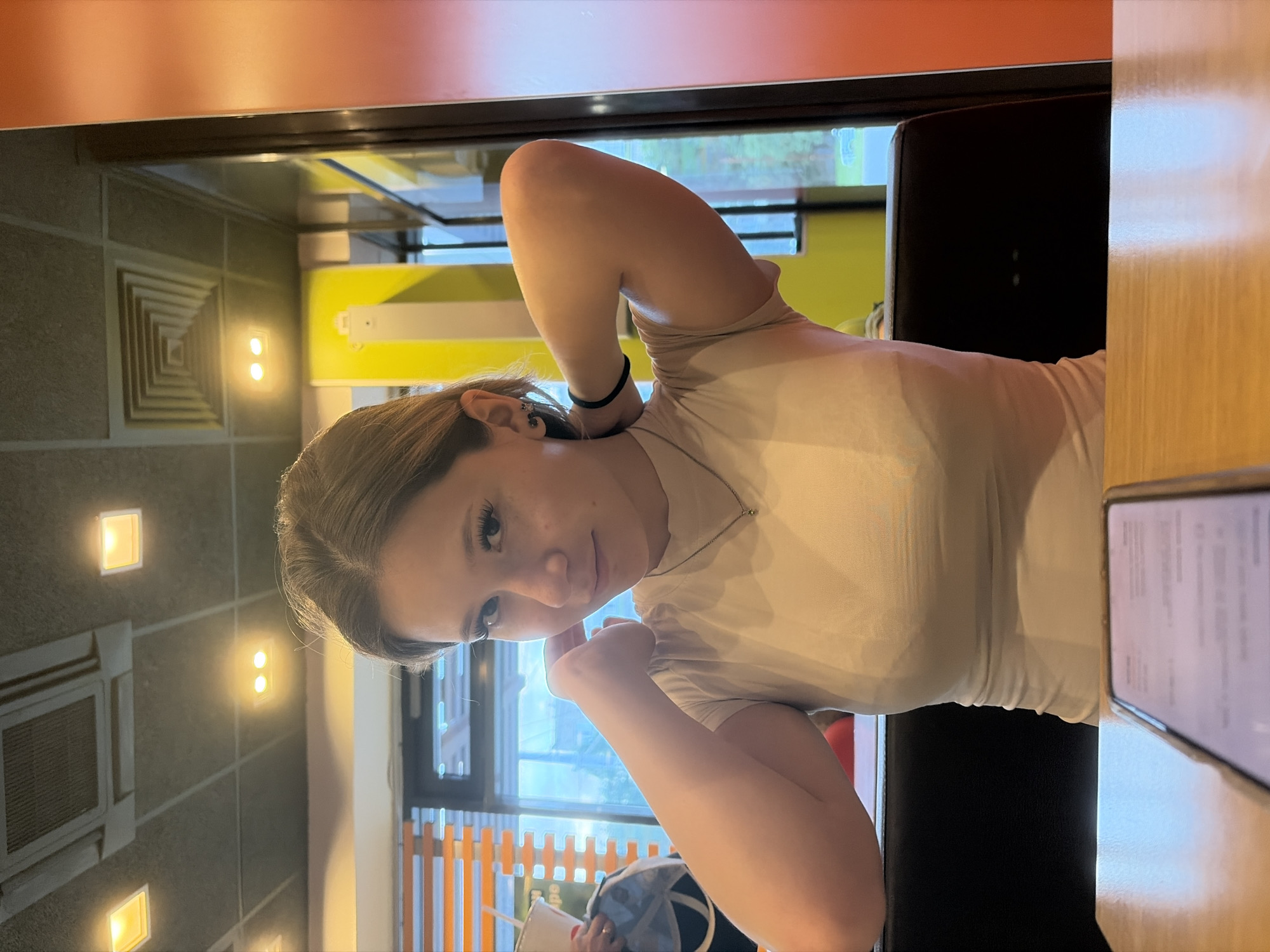 A young Caucasian woman with blonde hair pulled back is seated at a table indoors. She is wearing a beige t-shirt and a silver necklace with a green pendant. Her left arm is bent and her hand is near her face, while her right arm is behind her head, holding her hair. She is looking at the camera with a slight smile. In front of her, on the table, is a smartphone with a blurred screen. The background shows a window with orange blinds, a yellow wall, and ceiling lights. The overall setting appears to be a cafe or restaurant.