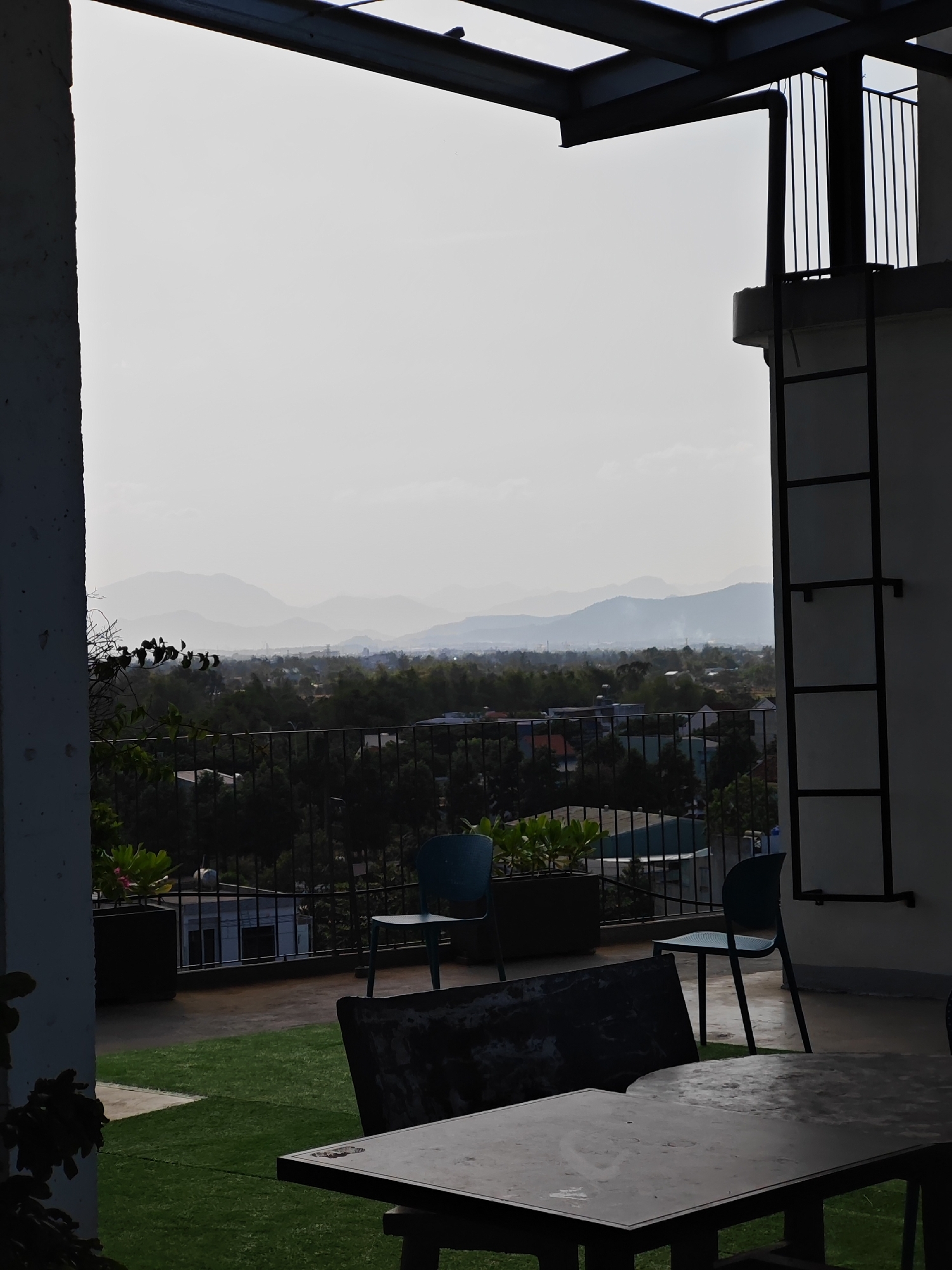 The image captures a rooftop view in Đà Nẵng, Vietnam, looking out towards a hazy landscape of distant mountains. The foreground features a dark, weathered table and two chairs, one teal and one dark, positioned on a patch of artificial turf. Beyond this seating area, a black metal railing encloses the rooftop, with a glimpse of buildings and lush greenery below. To the right, a white wall is adorned with a tall, black metal ladder. The sky is overcast and pale, with the soft light suggesting it is either early morning or late afternoon. No people are visible in the scene.