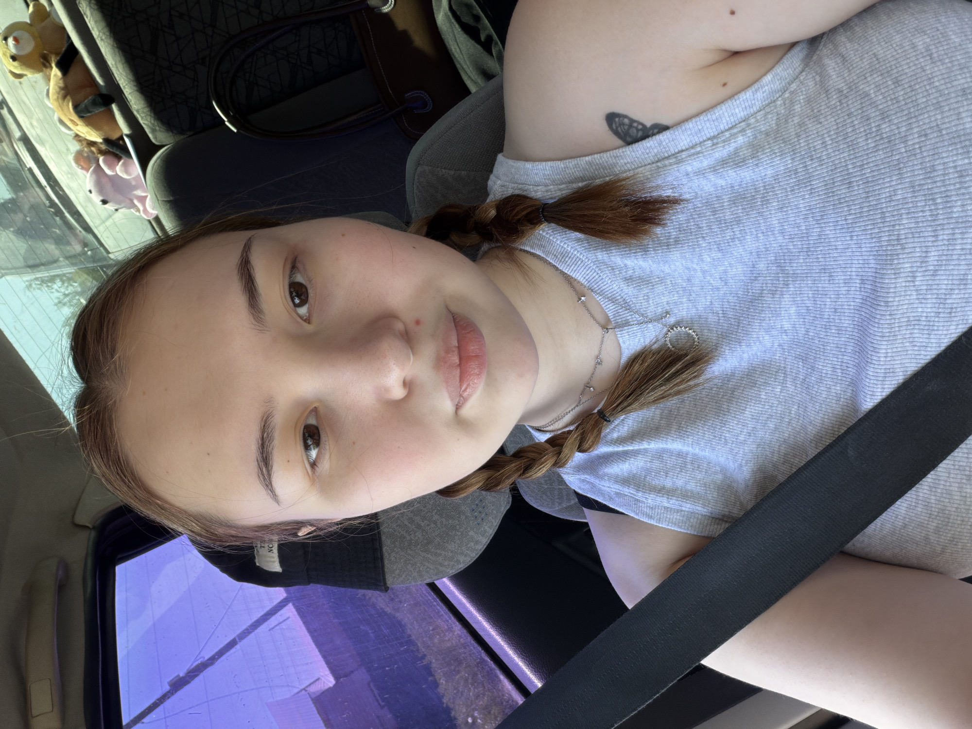 A young woman with braided auburn hair is seated in a vehicle, wearing a light grey shirt and a seatbelt. A silver necklace adorns her neck. A butterfly tattoo is visible on her left shoulder. Plush toys are visible on the back seat. The location cannot be determined to be Chaikovsky, Russia. The scene is lit by natural daylight.