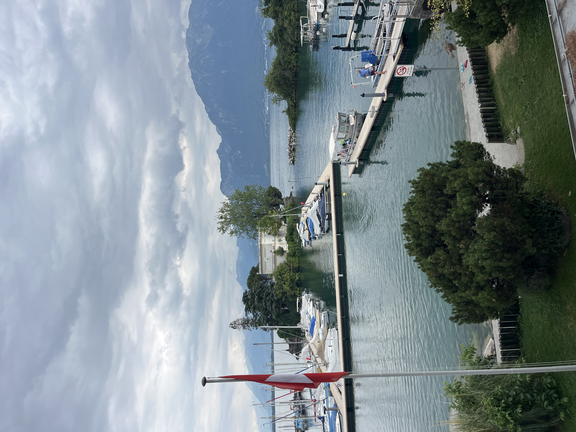 This image captures a view of a marina in Montreux, Switzerland, on a cloudy day. In the foreground, a Swiss flag flies from a flagpole on the left. A paved walkway borders the grassy area in the lower part of the frame, with several pairs of shoes scattered on the walkway near a sign prohibiting diving. Wooden posts line the edge of the walkway.
The marina itself is filled with various boats, including sailboats and smaller motorboats, docked along wooden piers. The water in the marina is calm and appears a deep turquoise color. In the middle ground, a small white building with columns sits amidst lush green trees on a small peninsula.
The background is dominated by a large, calm lake, likely Lake Geneva, with a distant mountain range under a heavy, overcast sky. The mountains are dark and rugged, adding a dramatic backdrop to the scene. The overall atmosphere is serene, despite the overcast weather.