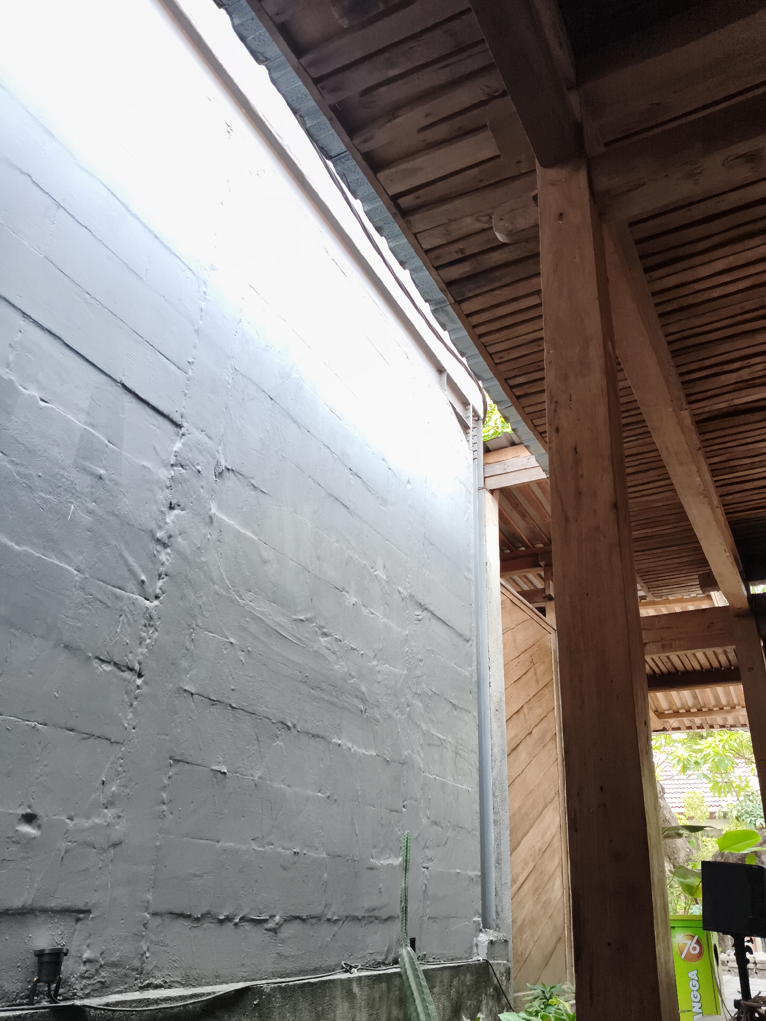 The image depicts an outdoor scene, likely in Nologaten, Indonesia, showcasing a textured grey wall alongside a wooden-beamed structure. The grey wall, painted with a rough finish, occupies the left portion of the frame, with subtle horizontal lines suggesting underlying construction. A drainage pipe runs vertically along the wall. To the right, a series of wooden beams and planks form a roof structure, supported by a substantial wooden post. The roof appears to be constructed from rough-hewn timber. In the background, glimpses of vegetation and roofing tiles suggest a residential or semi-rural environment. The lighting indicates daytime, with a bright, diffused light illuminating the wall. There are no people visible in the image.