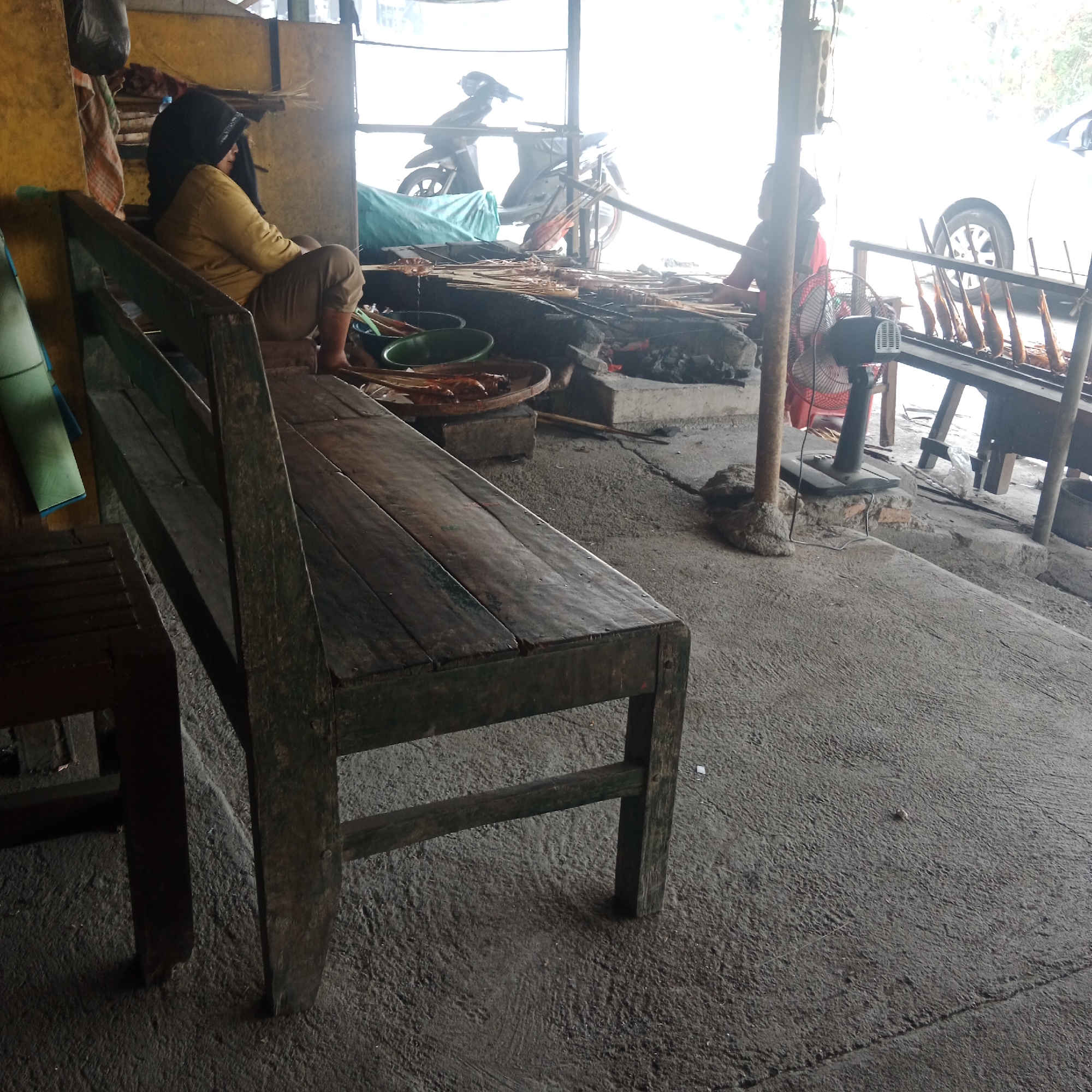 In the city of Ngandul, Indonesia, a modest outdoor food stall is depicted. A person wearing a yellow shirt and black headscarf is seated on a wooden bench to the left, appearing to be taking a break or observing. In the background, a motorcycle is parked, and another person in a red garment is tending to food being grilled over charcoal. Several skewers of what appear to be fish or meat are arranged on racks above the fire, indicating a grilling operation. A fan is positioned on the ground near the grilling area. The ground is rough concrete, and the stall is partially covered by a corrugated metal roof and wooden beams. The scene suggests a typical street food vendor setup.