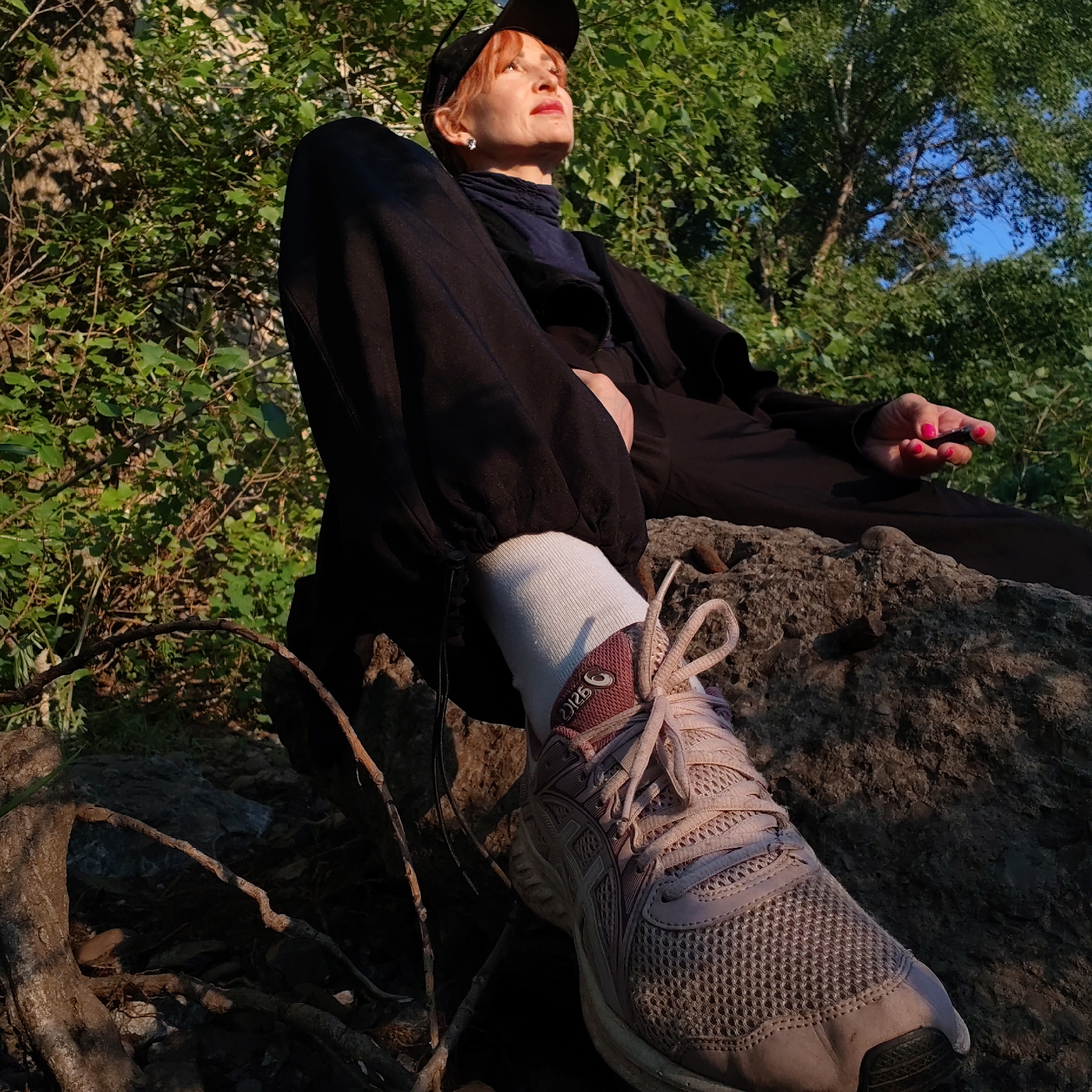 The image portrays a woman seated outdoors in a natural, sunlit environment, likely a park or forest edge, in Chaikovsky, Russia. The shot is captured from a low angle, looking upwards towards the subject and the sky.
The woman, with reddish-orange hair and red lipstick, is wearing a black baseball cap, a dark blue high-collared top or scarf, a black jacket, and black loose-fitting pants. She has small earrings. Her white socks and light pinkish-beige Asics sneakers are prominent, with her right foot resting on a textured rock. Her left hand, adorned with bright pink fingernail polish, is casually placed on her lap. She is gazing upwards with a calm and contemplative expression, illuminated by warm, directional sunlight.
The setting is characterized by large, dark rocks and visible tree roots on which she is resting. The background features abundant green foliage and trees, with patches of clear blue sky visible through the canopy. The strong contrast between light and shadow, and the golden quality of the sunlight, suggests either early morning or late afternoon, often referred to as the golden hour.
The brand name "ASICS" and a stylized number that appears to be "2126" are clearly visible on the side of her right sneaker. The overall scene conveys a sense of tranquility and connection with nature.