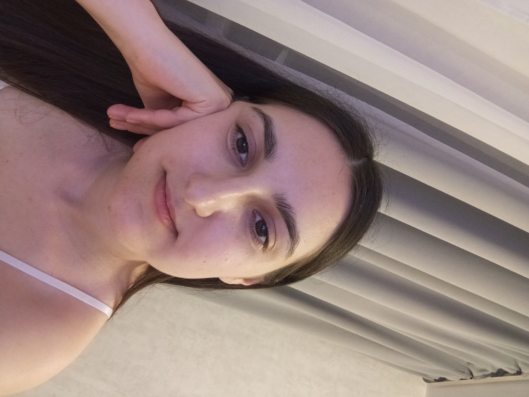 A close-up, waist-up, indoor selfie of a young adult female with dark hair, leaning her head on her hand and looking towards the camera. She is wearing a light-colored spaghetti strap top. The background consists of vertical, light-colored curtains and a textured wall. There are no identifiable landmarks or indications of the city of Krasnodar, Russia.