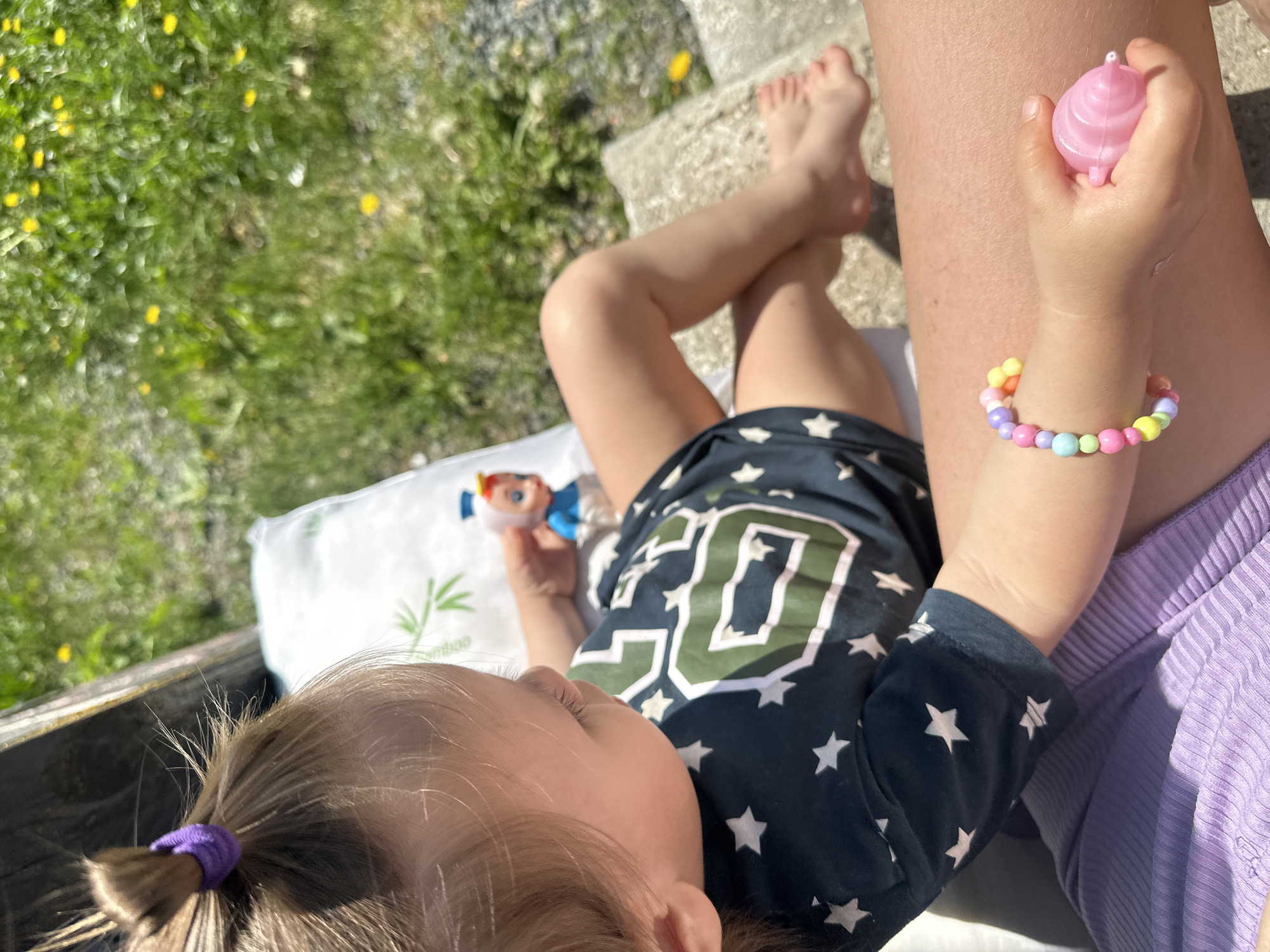 A young child is lying on a white pillow outdoors, possibly in a garden or patio area. The child is wearing a navy blue t-shirt with white stars and the number "30", and lilac colored bottoms. They have a ponytail secured with a purple hair tie. The child is holding a pink toy in one hand and has a colorful beaded bracelet on the same wrist. There is a doll lying on the pillow next to the child. The child's feet are visible to the right of the image. The setting includes grass, yellow flowers, and stone steps or an edge. It is sunny. There is no indication of the city of Kharnavy, Russia.