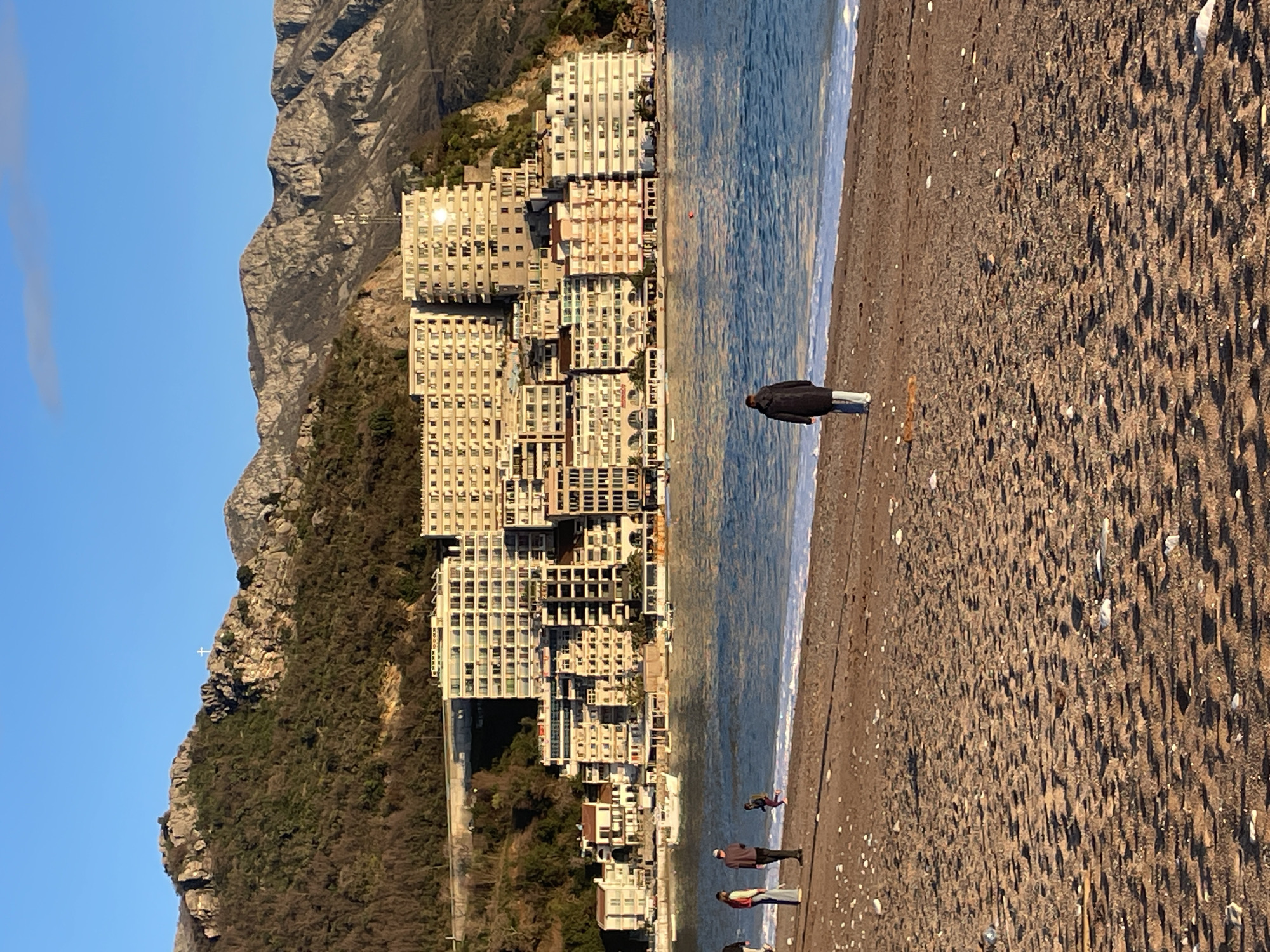 The image depicts a coastal scene in Rafailovići, Montenegro. In the foreground, a sandy beach stretches across the frame, populated by several individuals. A person in a dark coat and light pants walks away from the camera, positioned centrally near the water's edge, where white foam from gentle waves is visible. To the left, two adults stand near each other, while a smaller individual in dark clothing runs or plays closer to the water. Another person is partially visible on the far left. The sand shows numerous small shadows and scattered light-colored pebbles/shells.
The midground features the calm, blue waters of the sea, reflecting the sky and exhibiting minor ripples. Several small boats are moored in the water closer to the shore on the left side.
In the background, a densely built-up coastal town rises from the water's edge and extends up a verdant hillside. Numerous multi-story buildings, primarily light-colored and featuring balconies, are clustered together. Many facades reflect the golden sunlight, creating bright spots. Beyond the town, rugged mountains covered with sparse vegetation and exposed rock dominate the horizon under a clear blue sky. A distinct white cross is visible atop one of the mountain peaks on the left. Long shadows are cast across the landscape, consistent with late afternoon or early morning sunlight.