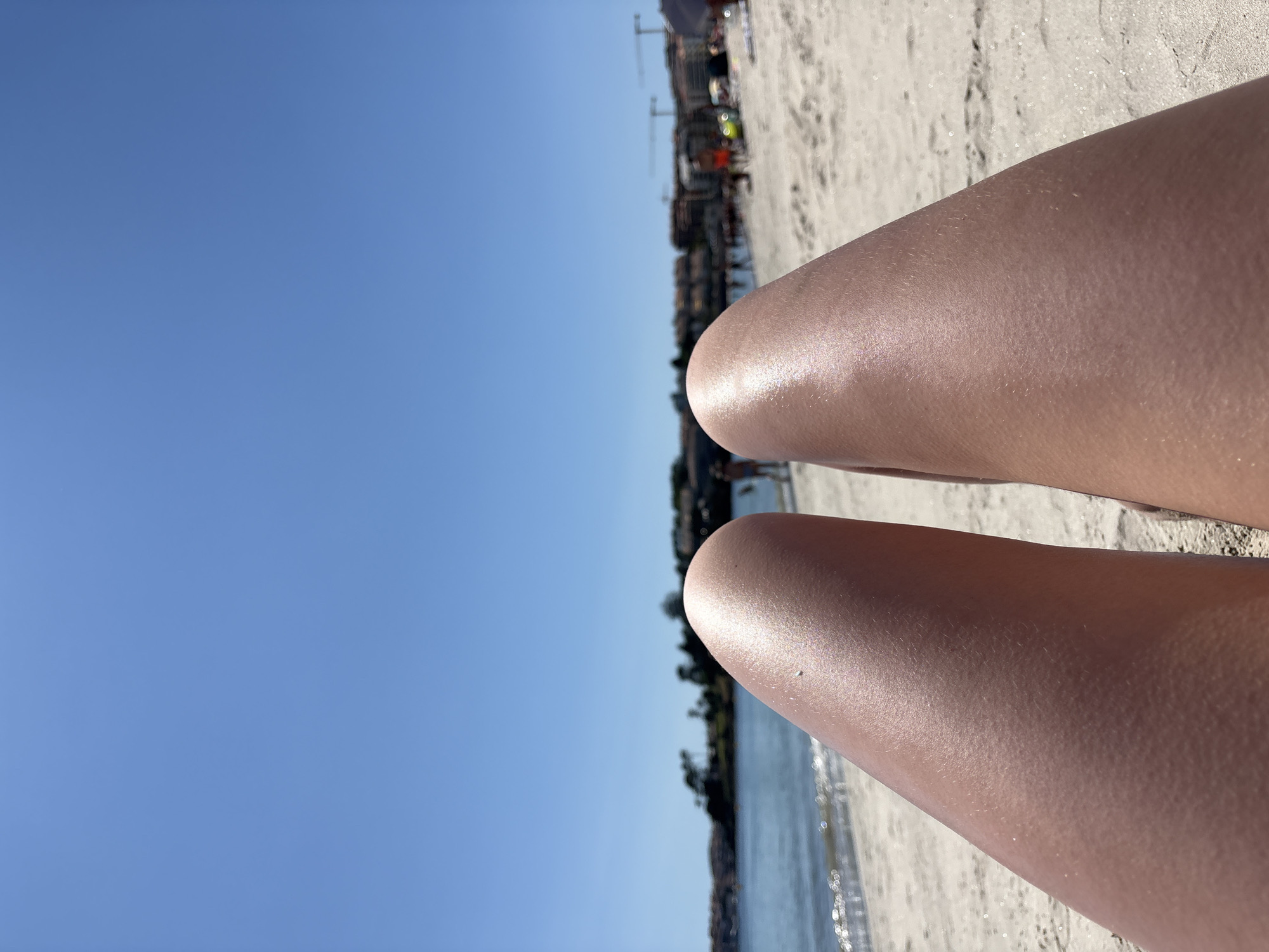The image depicts a first-person perspective of a person's legs resting on a sandy beach with the ocean and a city skyline in the background. The sky is clear and blue, indicating a sunny day. The water of the ocean is calm, with gentle waves reaching the shore. In the distance, buildings and trees line the coast, suggesting an urban or semi-urban environment. There are a few blurry figures of people visible further down the beach and in the water, engaged in various activities. The beach sand is light in color and appears to be slightly damp near the water's edge. The scene is likely set in Sanxenxo, Spain, known for its beaches and coastal attractions.