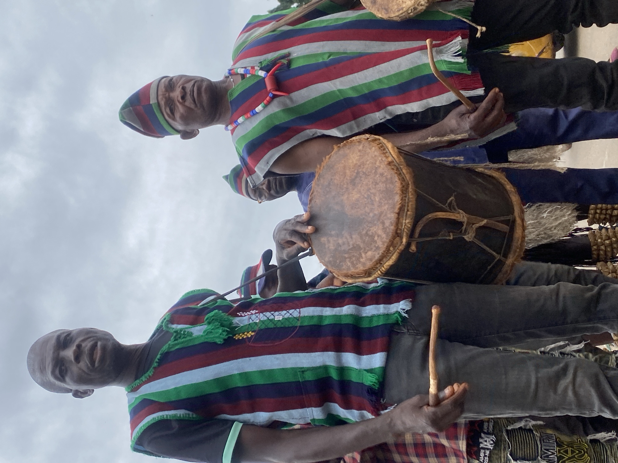 Two men in Maigizo, Nigeria stand outdoors playing drums. Both are wearing striped, fringed tops and each holds a drumstick. One man wears a matching striped hat and a beaded necklace. The sky is overcast.
