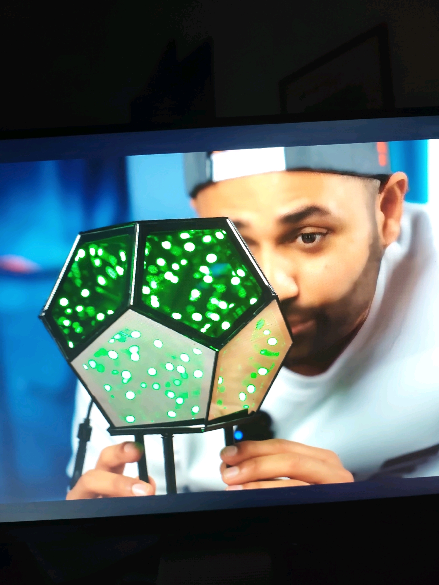 The image shows a close-up of a man wearing a baseball cap, holding a geometric object. The object is a dodecahedron-like shape with green translucent panels illuminated by white lights, creating a glowing effect. The man is looking towards the object, with his face partially obscured by it. The background is blurred, with shades of blue and hints of a darker area, possibly a room or studio. There is no discernible information within the image to indicate the location in Minde, Portugal.