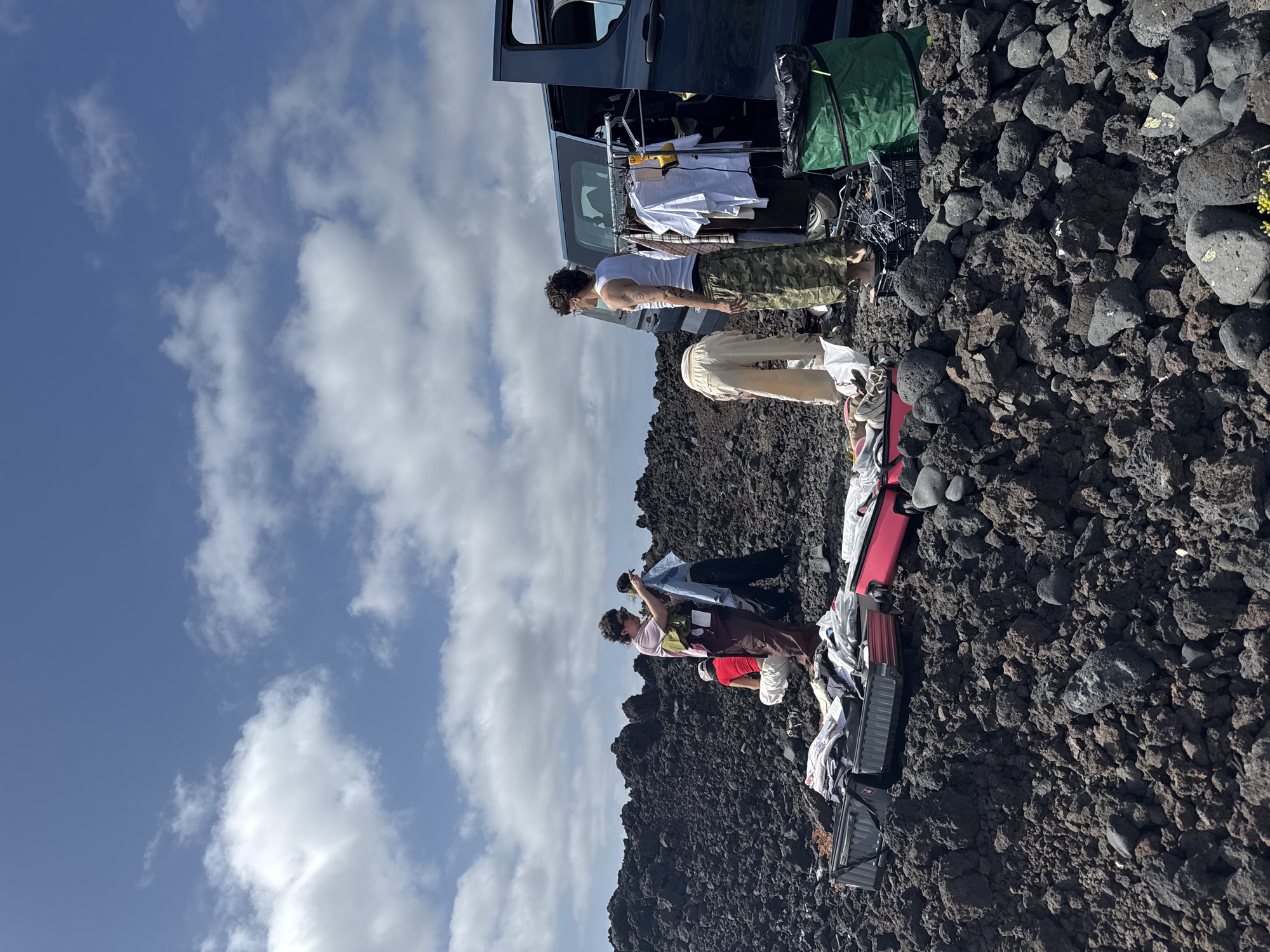 The image shows a scene in a rocky, lava field landscape under a sky with scattered clouds. The location appears to be in Tinajo, Spain, given the volcanic terrain typical of the Canary Islands.
A dark-colored van with its back doors open is visible. Clothing hangs on a rack inside the van. Two people stand near the van, while two others are positioned further down the slope among luggage and other items.
The ground is covered with dark volcanic rock. Clothing and luggage are scattered on the ground. The overall impression is that of people unloading or organizing belongings near a vehicle in a volcanic landscape.