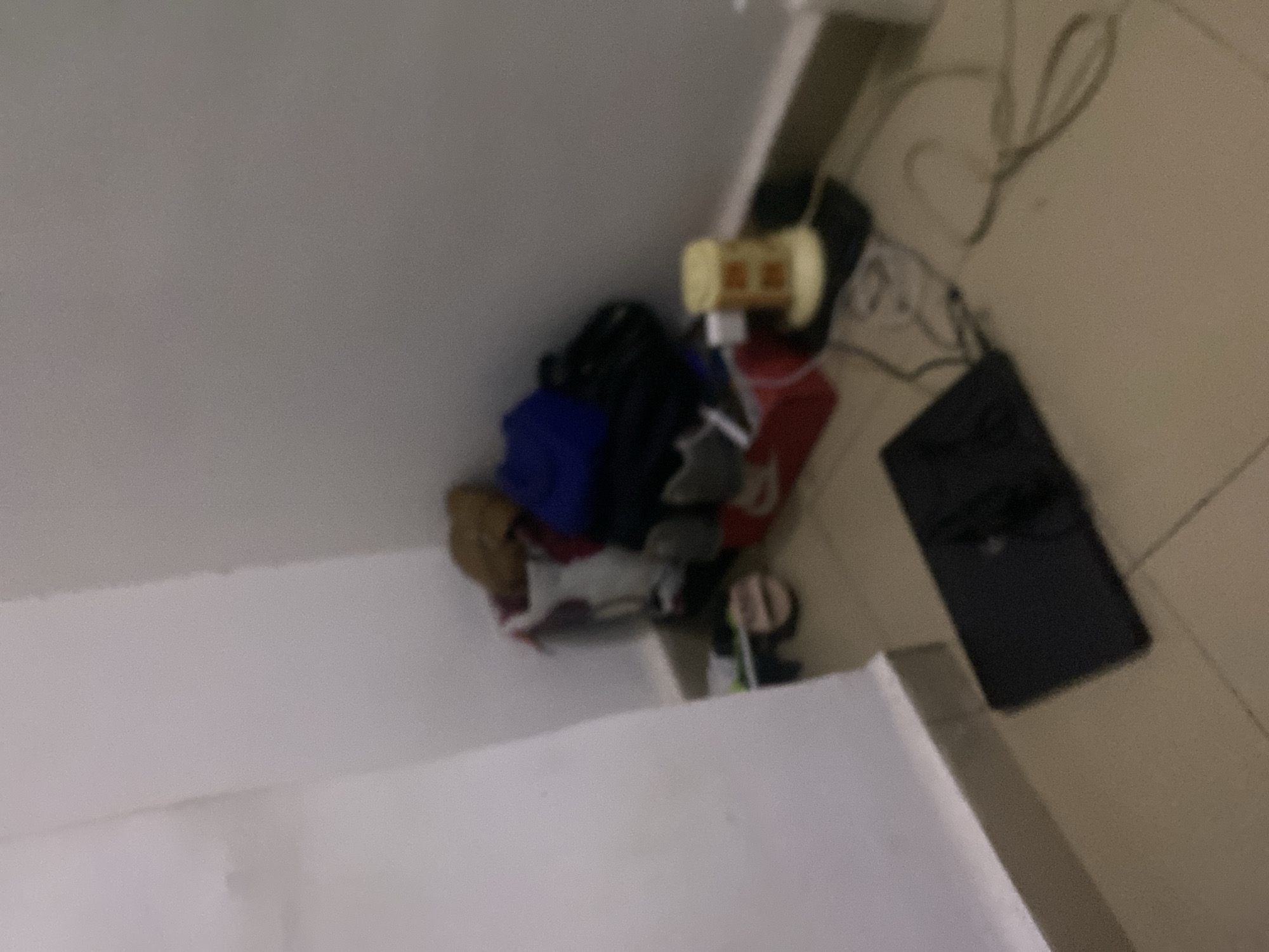 The image depicts a cluttered corner of a room, likely a bedroom or storage area, in what is described as Howayi, Nigeria. In the corner, a pile of items includes clothing such as a brown jacket, a blue shirt, and other garments, some of which appear to be in bags or boxes. A red plastic container is also visible. On the floor in front of this pile, a black tablet-like device is present, with some cables nearby. To the right of the pile, a yellow object with a grid pattern sits on a black surface, likely an adapter or small appliance. The floor is tiled in a light beige color, and the walls are painted white. The lighting is dim, creating shadows and a generally low-visibility environment. There are no people or animals visible in the image.