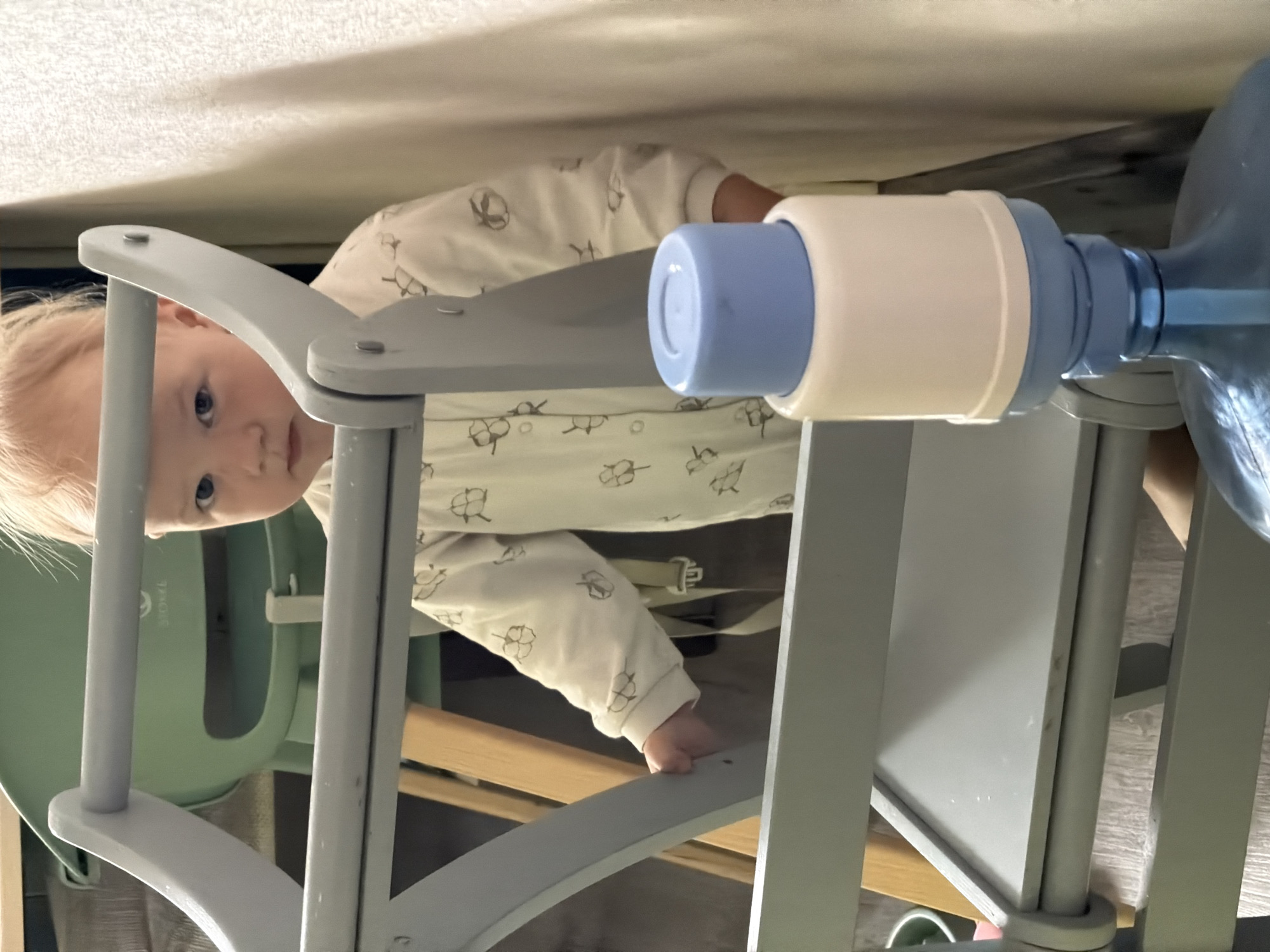A young child is standing behind a grey wooden high chair and looking at the camera. The child is wearing a white onesie with a pattern of small flowers. The child's hair is light brown and appears to be thinning. To the right of the child, a large blue water bottle with a white and blue dispenser is visible. The background consists of a pale yellow wall and a light green high chair. The setting appears to be indoors, possibly a kitchen or dining area, in a residential location in Chaikovsky, Russia.