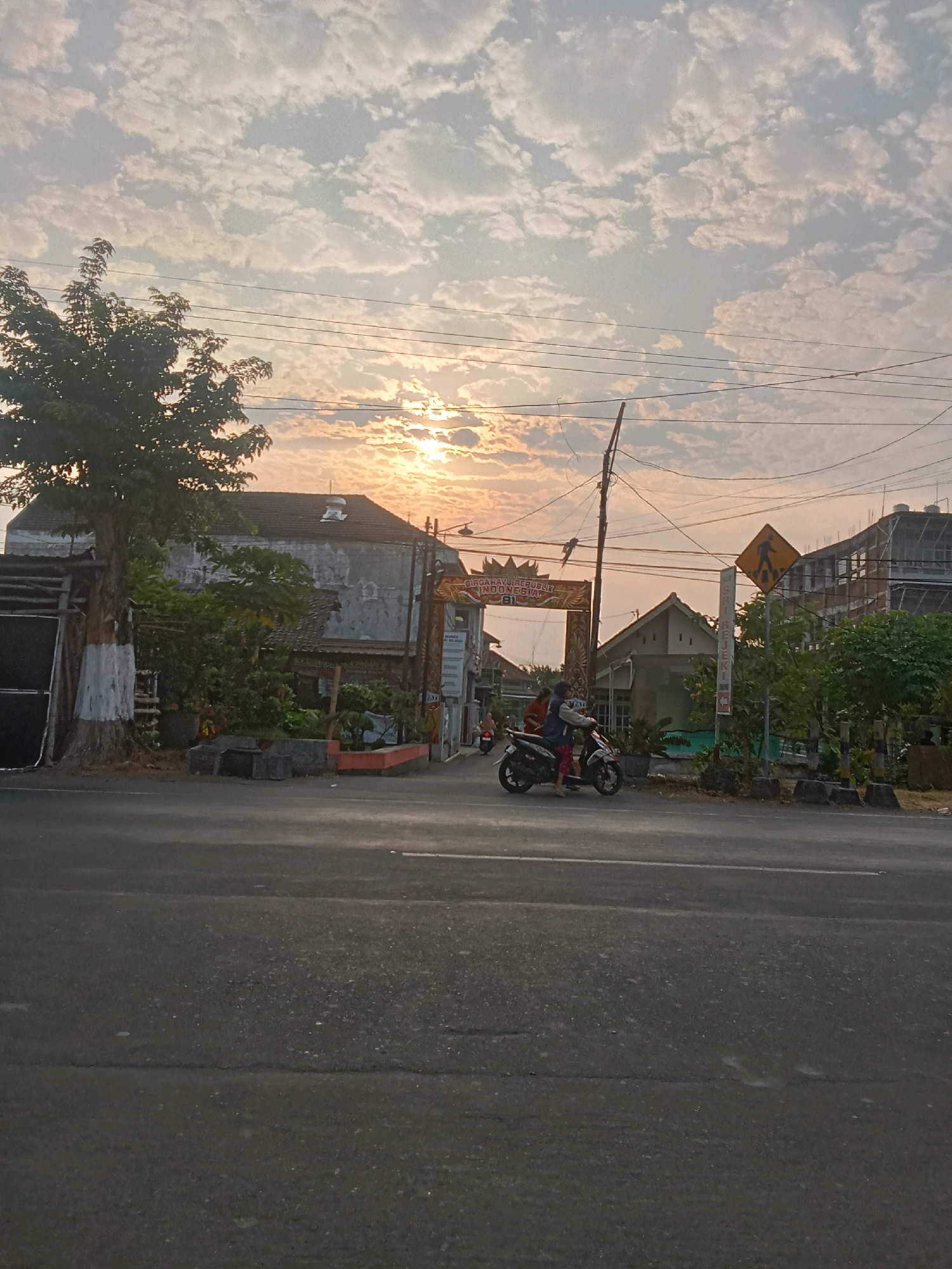 This is a photograph taken in Kaliwungu, Indonesia, capturing a street scene at sunrise. The sky is filled with scattered clouds, illuminated by the rising sun, creating a warm, golden glow. In the foreground, a paved road stretches across the frame, with a white line marking the lane. On the right side of the road, a yellow diamond-shaped pedestrian crossing sign is visible, accompanied by a vertical sign with text. Further back, a building with a dark roof and weathered walls stands partially obscured by trees and foliage. A large tree with a white-banded trunk is on the left side of the image. In the middle ground, a motorcycle with two riders is moving along the road. Behind the motorcycle, an archway with text and decorative elements is visible, suggesting it marks an entrance or a significant point in the town. Several power lines crisscross the sky. The overall impression is of a typical morning in a residential or commercial area of Kaliwungu.