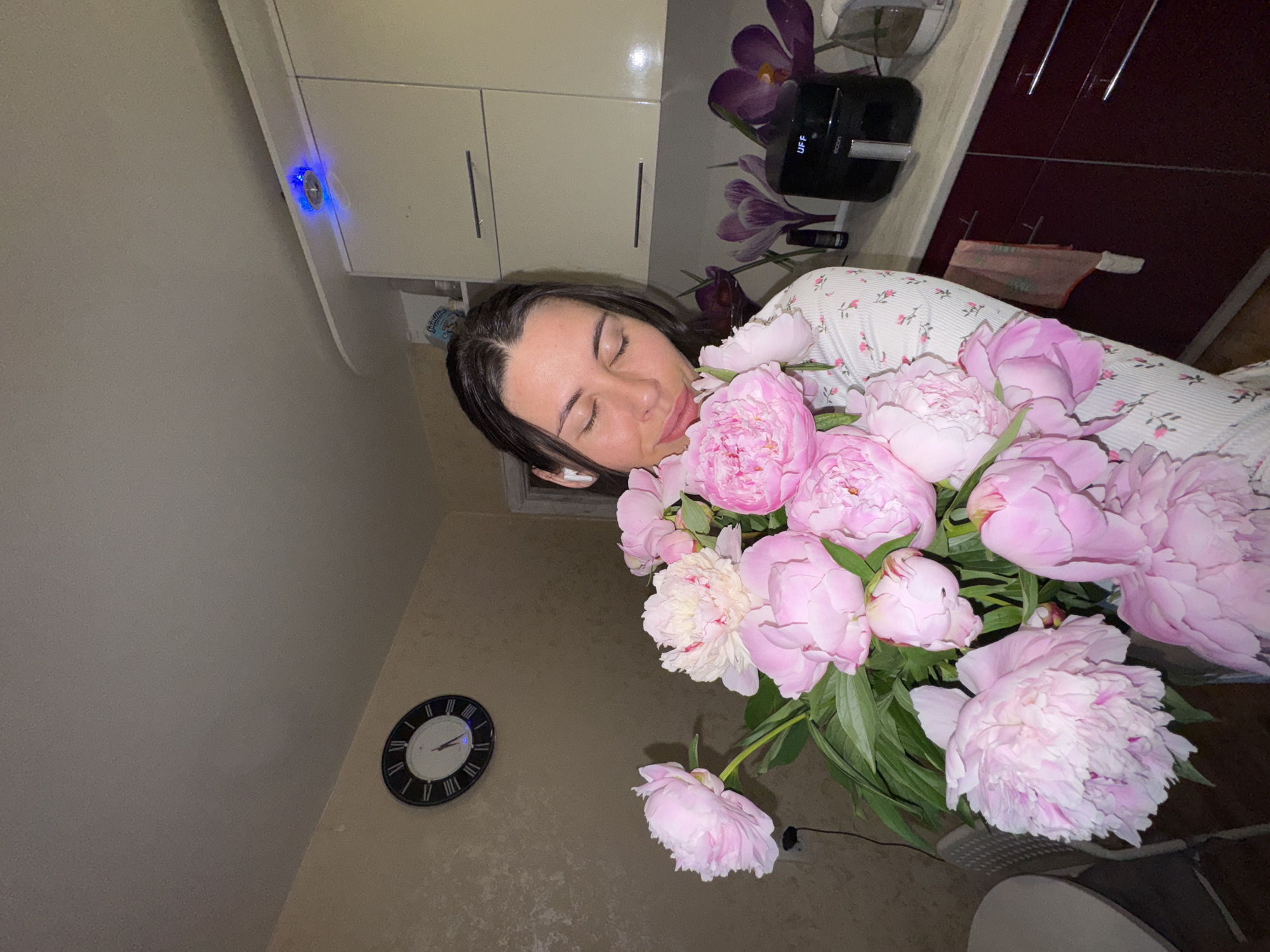 A young woman is holding a large bouquet of pink peonies in what appears to be a kitchen. She is wearing a white pajama top with a floral print and white earbuds. Her eyes are closed and she is smiling gently as she sniffs the flowers. In the background, there is a kitchen counter with an air fryer, some decorative purple flowers, and cabinetry. A round clock with Roman numerals is visible on the wall. The overall scene suggests a domestic and peaceful moment. The image does not provide any information about the city of Chaikovsky, Russia.