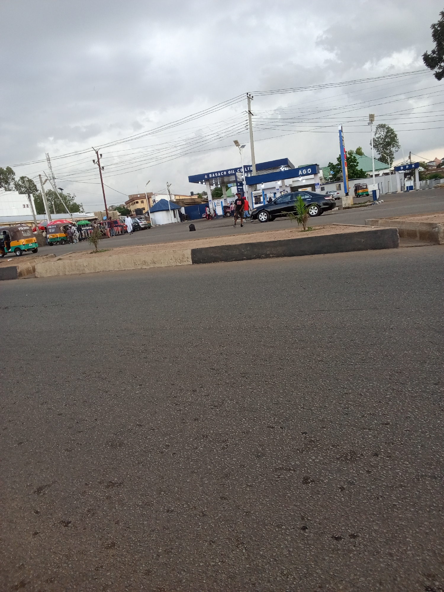 The image shows a street scene in Jos, Nigeria, with a gas station in the background. The gas station is labeled "BASACO OIL CO LTD" and "AGO". There are several vehicles at the gas station, including a black sedan, a trishaw, and other cars. A few people are visible walking around the gas station and near the vehicles. The sky is overcast with grey clouds. The foreground of the image shows a paved road. The overall scene depicts a typical day at a gas station in an urban or semi-urban environment.
