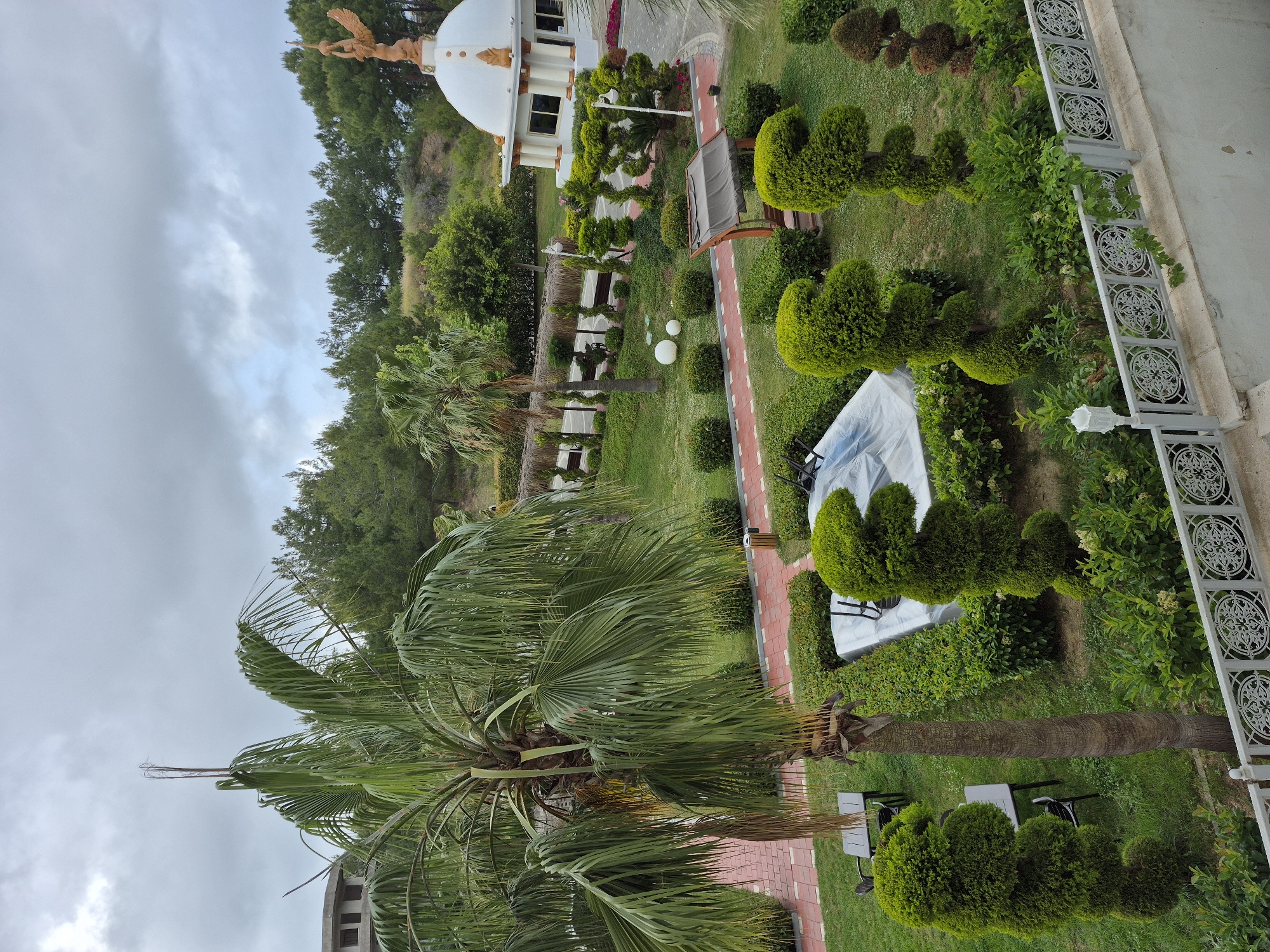 The image depicts a landscaped garden or park, likely located in the city of Alanya, Turkey. The setting is outdoors with lush green vegetation, including palm trees and manicured bushes. A red-brick pathway weaves through the garden. Some elements include a building with a white dome topped with a statue and several structures covered with white fabric. There are tables and chairs in the scene, possibly for seating. The overall impression is of a well-maintained and aesthetically pleasing environment. The sky is overcast.