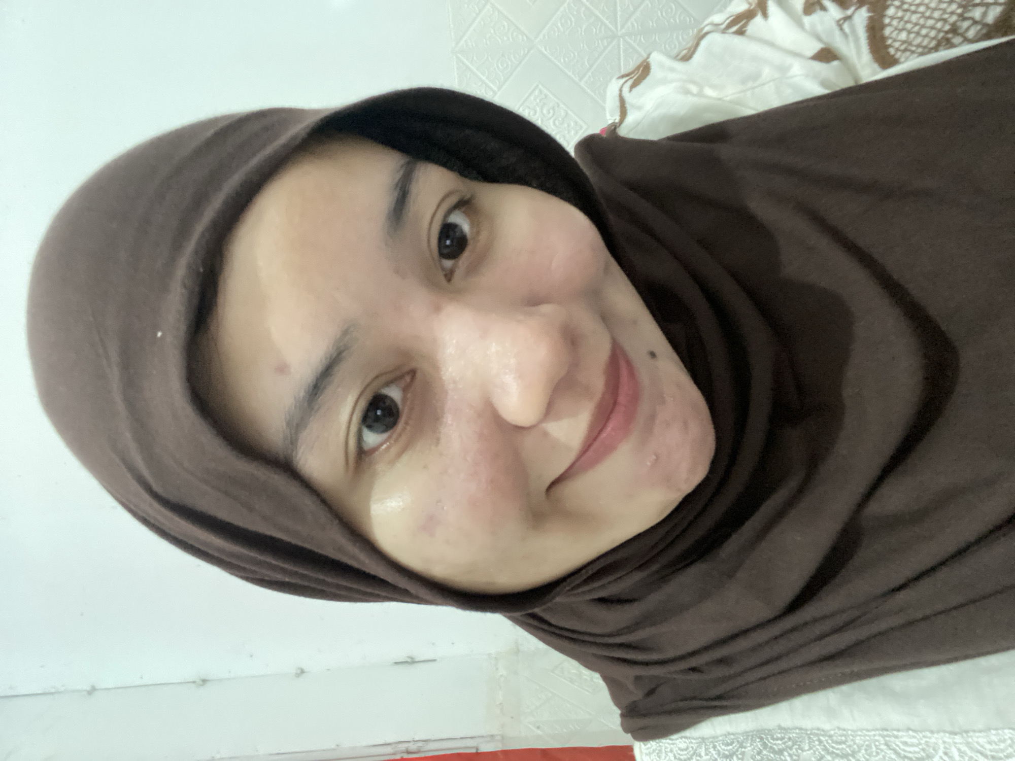 A young Indonesian woman wearing a brown hijab and a white garment with a brown embroidered pattern smiles at the camera. She is indoors, with a textured white wall in the background. The image is a close-up selfie.