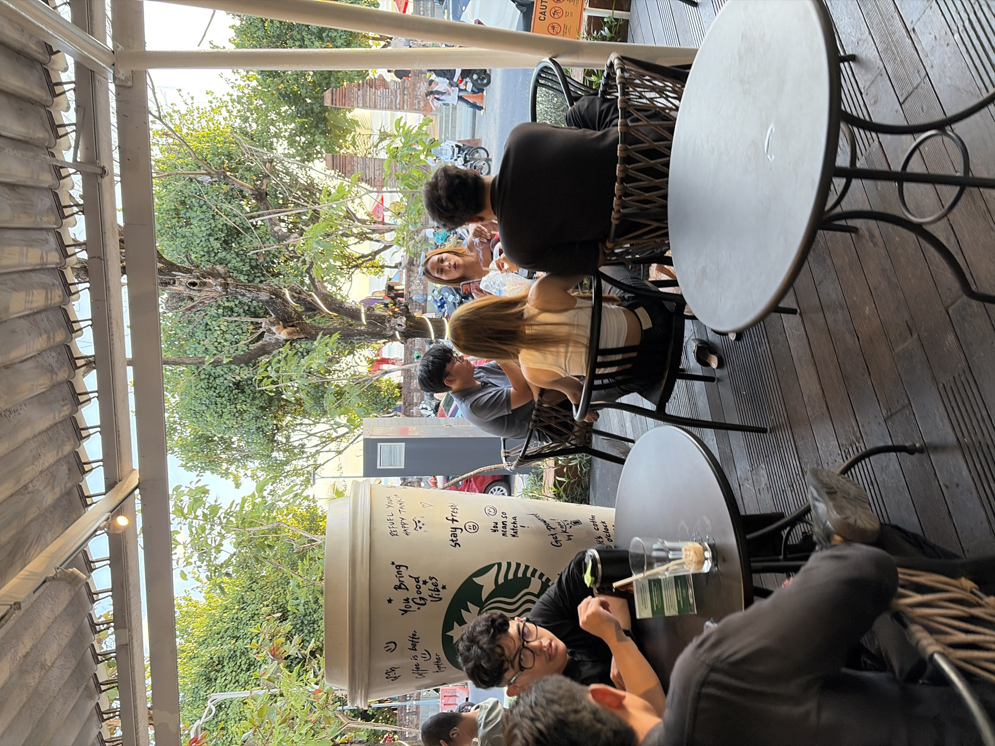 The image depicts an outdoor seating area of a Starbucks cafe in Kuta, Indonesia. A large, decorative Starbucks cup is prominently displayed on the left side of the frame. Several people are seated at tables, some conversing and others appearing to be engaged with their phones or drinks. In the background, a street scene is visible with motorcycles and pedestrians, suggesting a busy urban environment. Lush green trees and foliage frame the outdoor seating area, indicating a tropical setting. The overall atmosphere appears relaxed and casual, typical of a cafe environment.