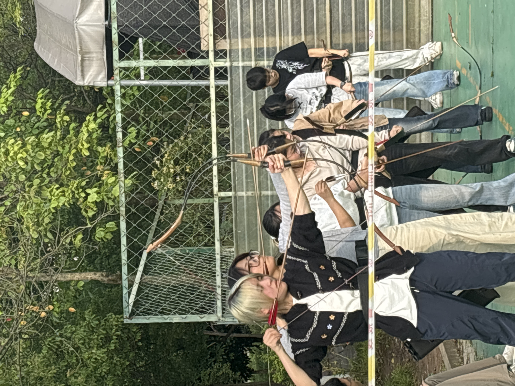A group of people stand in a line outdoors, holding bows and arrows, presumably participating in archery. They appear to be aiming or preparing to shoot. The background features a fence, trees, and a covered structure. There is a yellow and white striped marker in the line of sight. The image provides no information about the specific location in Jiangjin, China.
