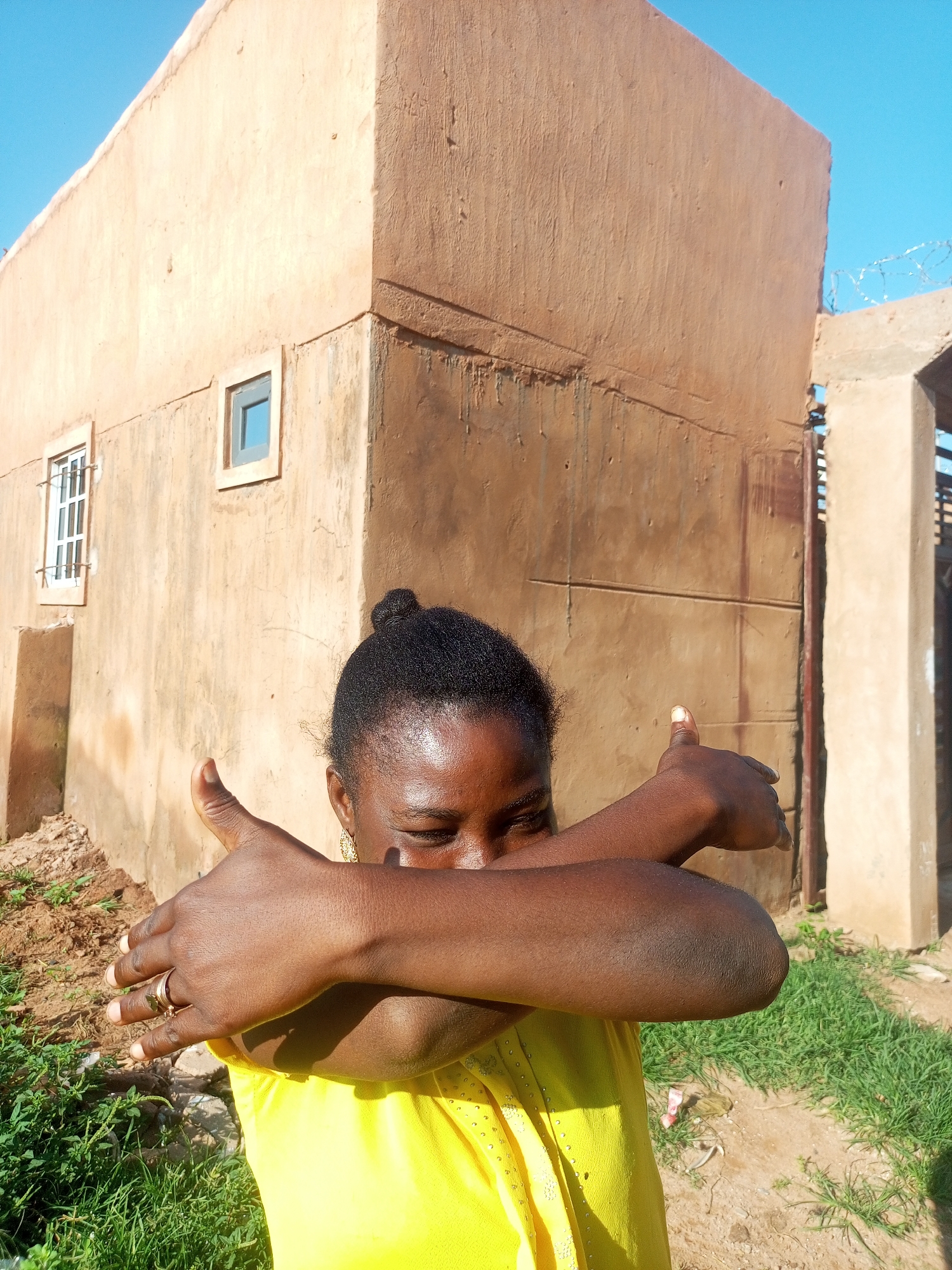 A medium shot captures a Black female with dark brown skin, her eyes closed and face partially obscured by her arms crossed in front of her. She is wearing a bright yellow t-shirt adorned with small, scattered embellishments. Her hair is pulled back into a neat bun on top of her head. One hand, adorned with a gold-toned ring, is visible resting on her bicep. She is positioned outdoors in front of a tan, stucco-clad building with a small, recessed window on the left wall and another on a section of the building on the right. The ground is covered in patches of dry dirt and green grass. The background features a clear blue sky and barbed wire atop a concrete structure. The scene appears to be located in Tempure, Nigeria.