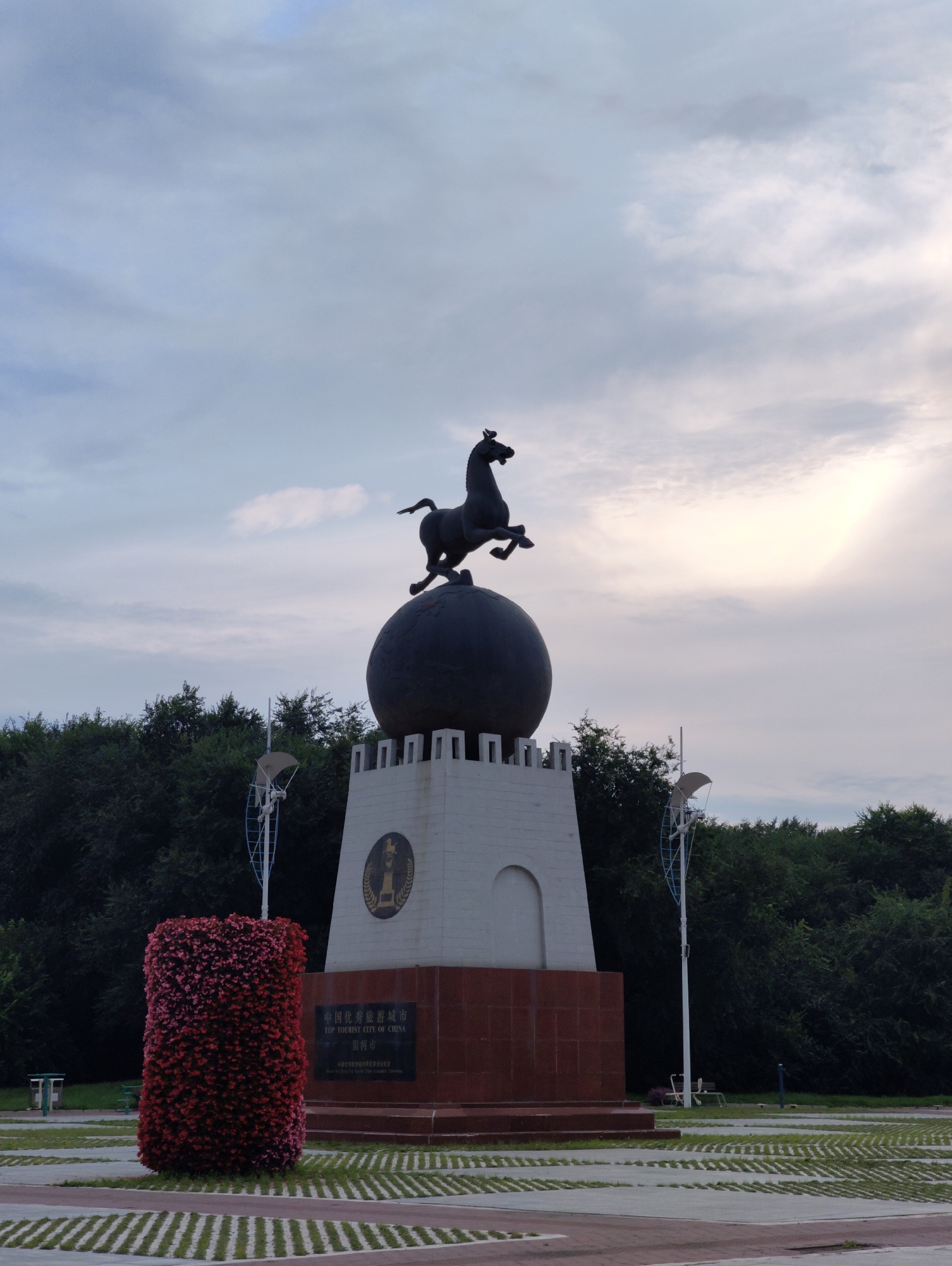 The image shows a monument with a statue of a horse atop a globe, which is itself placed on a pedestal. The pedestal is a white, multi-tiered structure with a darker, red base. The monument is situated in an open area with a paved ground covered in a pattern of grass and paving stones. To the left of the monument is a large, rectangular floral arrangement filled with red flowers. In the background, a dense line of dark green trees forms a backdrop, and the sky above is overcast with visible clouds. Two lampposts with what appear to be small wind turbines at the top are also present near the monument. A plaque on the monument base bears Chinese characters and English text that reads "TOP TOURIST CITY OF CHINA" and below that "幸福乡". The scene appears to be a public space in the city of 幸福乡, China. There are no people visible in the image.