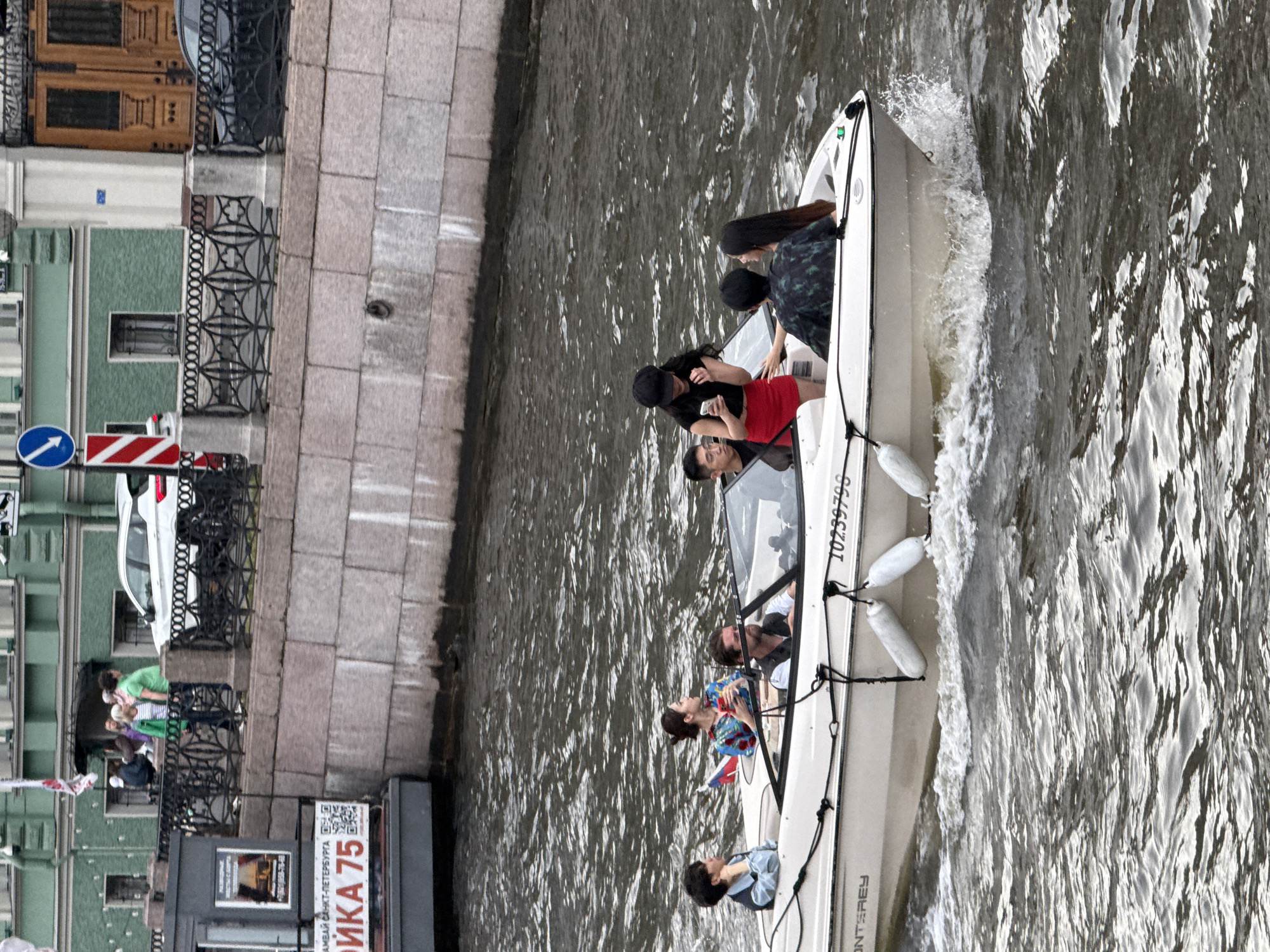 The image shows a small white motorboat with six people on board, navigating a canal in Saint Petersburg, Russia. The boat has the number "10259798" visible on its side. The canal is flanked by a stone embankment on one side and buildings on the other. The buildings appear to be historical, with the green building in the background possibly being part of an older architectural complex. On the embankment, there is a black metal fence and people walking along the top. A white car is parked near the fence. The water is choppy, indicating the boat is in motion.
The people on the boat are diverse in age and gender. In the front of the boat, a person with short dark hair is looking to the left. Next to them, a woman in a colorful top is looking down at her phone. A man with a beard is at the helm, steering the boat. Behind him, a person in a patterned shirt is facing away from the camera. Towards the back of the boat, a woman wearing a black cap and a red skirt is holding a phone and interacting with the person in the patterned shirt. Another person with long dark hair is seated beside them. A Russian flag is visible on the boat.
The overall atmosphere suggests a leisurely boat trip on a cloudy day in a city known for its canals and historical architecture.