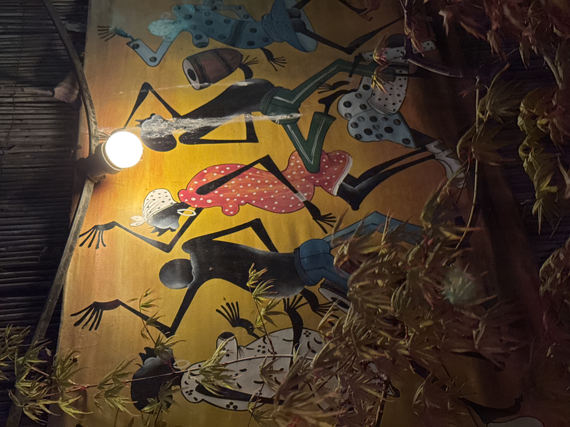 A vertical, eye-level shot depicts a mural illuminated by a single bare lightbulb on its left side. The mural, painted on a yellow-orange background, features stylized black figures with elongated limbs in various poses. Some figures are adorned with brightly colored clothing, including polka-dotted dresses in red, white, and blue. A drum is visible amidst the figures. Vegetation, including branches and leaves, partially obscures the right side of the mural. The lightbulb is mounted on a wooden structure adjacent to the mural. The location is Varna, Bulgaria.