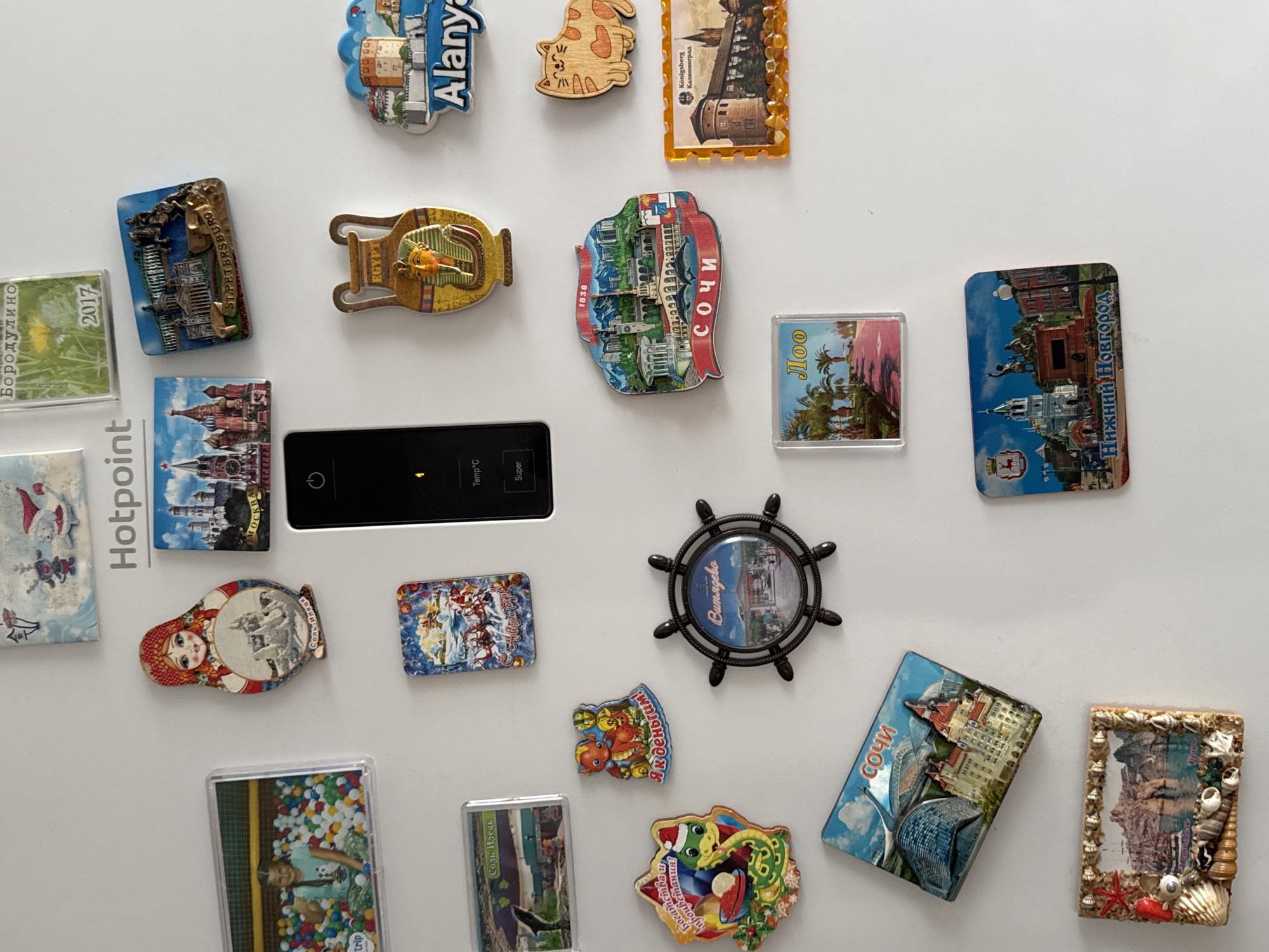 The image displays a white refrigerator door adorned with numerous colorful magnets. The magnets feature various tourist destinations and decorative designs. Among them, a magnet depicts a scene from Бородулино, Russia, showcasing a grassy field with a single yellow dandelion and the year "2017" prominently displayed. Other magnets include images of matryoshka dolls, famous landmarks like St. Petersburg and Moscow, Egyptian artifacts, and seaside resorts such as Sochi and Alanya. A magnetic frame filled with seashells and a coastal landscape is also visible. The overall scene is an arrangement of travel souvenirs on a domestic appliance.
