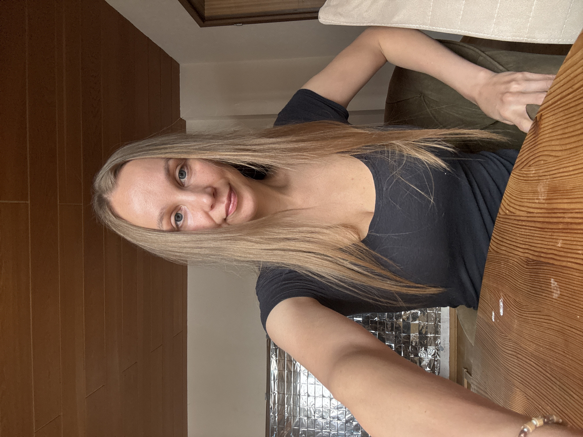 A medium shot captures a Caucasian woman with blonde hair and blue eyes from the chest up, seated at a wooden table. She is wearing a dark grey t-shirt. Her left arm is extended towards the camera, resting on the table, and she is wearing a bracelet on her wrist. Her right arm is bent, with her hand resting on the armrest of a dark green tufted chair. The background includes a wall with reflective square tiles, a wooden ceiling, and a doorway. The lighting appears to be natural, coming from the side, creating soft shadows. The scene is likely indoors, possibly in a residential setting in Überherrn, Germany, given the context.
