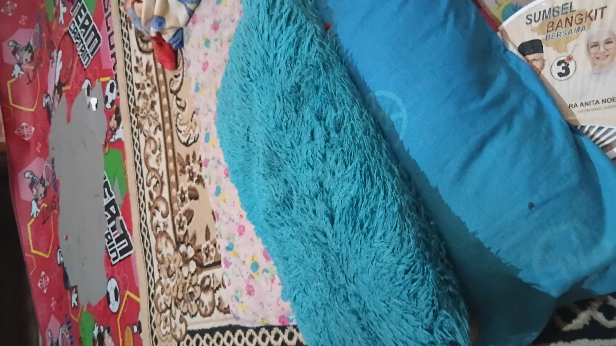 The image shows two pillows on what appears to be a bed. The top pillow is a fluffy, teal-colored cushion, while the bottom pillow is a solid teal color. A patterned blanket with cartoon characters is visible in the background. There is also a piece of paper with text and images of people. The scene suggests a domestic setting, likely a bedroom. There is no information in the image that allows for the determination of its location in Prabumulih, Indonesia.