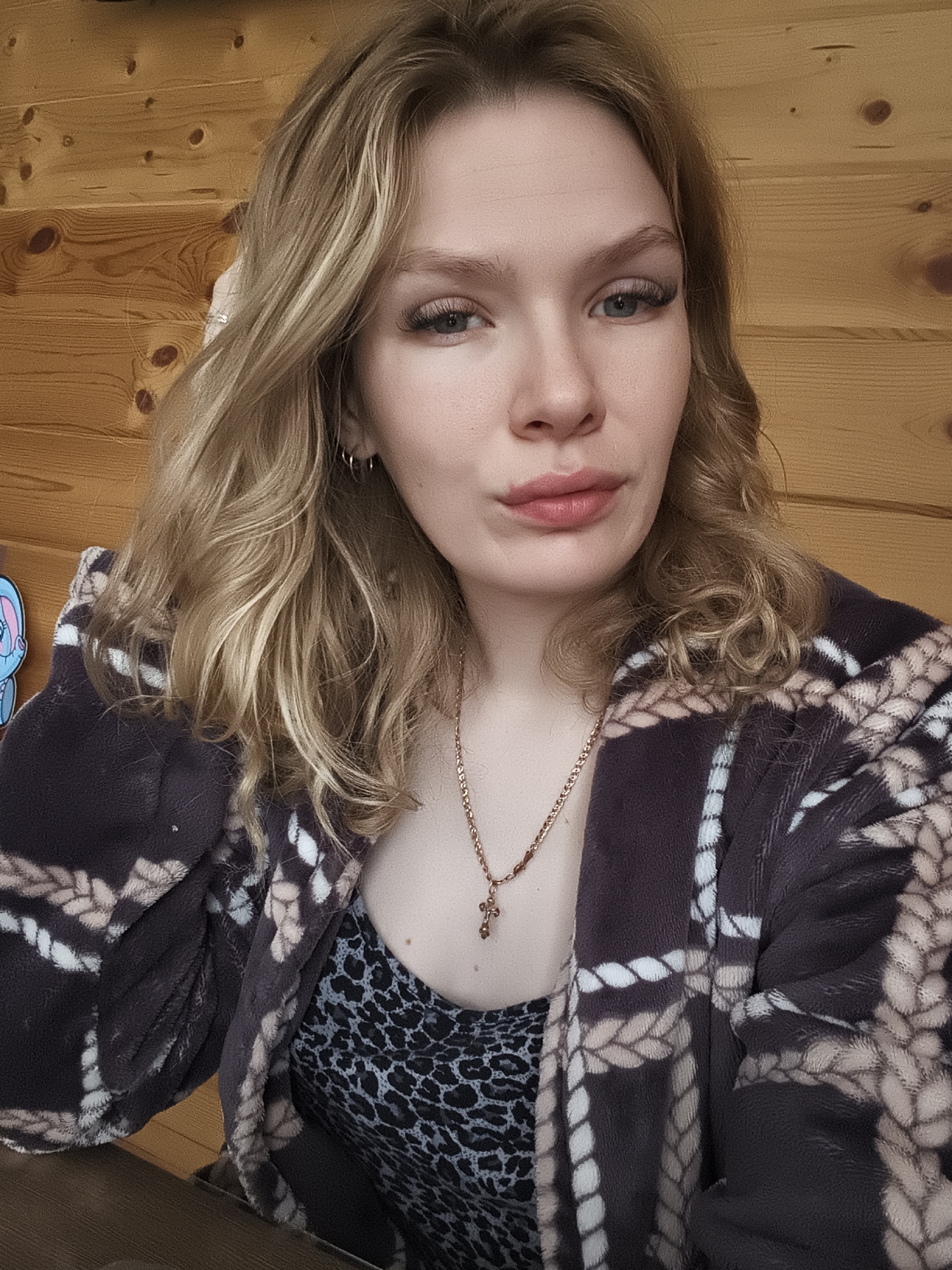 A woman is indoors, in front of a wooden wall. She has blonde hair, blue eyes, and pink lips. She is wearing a dark brown robe with a white rope pattern over a gray leopard-print shirt. She is also wearing a gold necklace with a cross pendant. Her left hand is partially visible near her hair. The location is implied to be Русский Вожой, Russia.