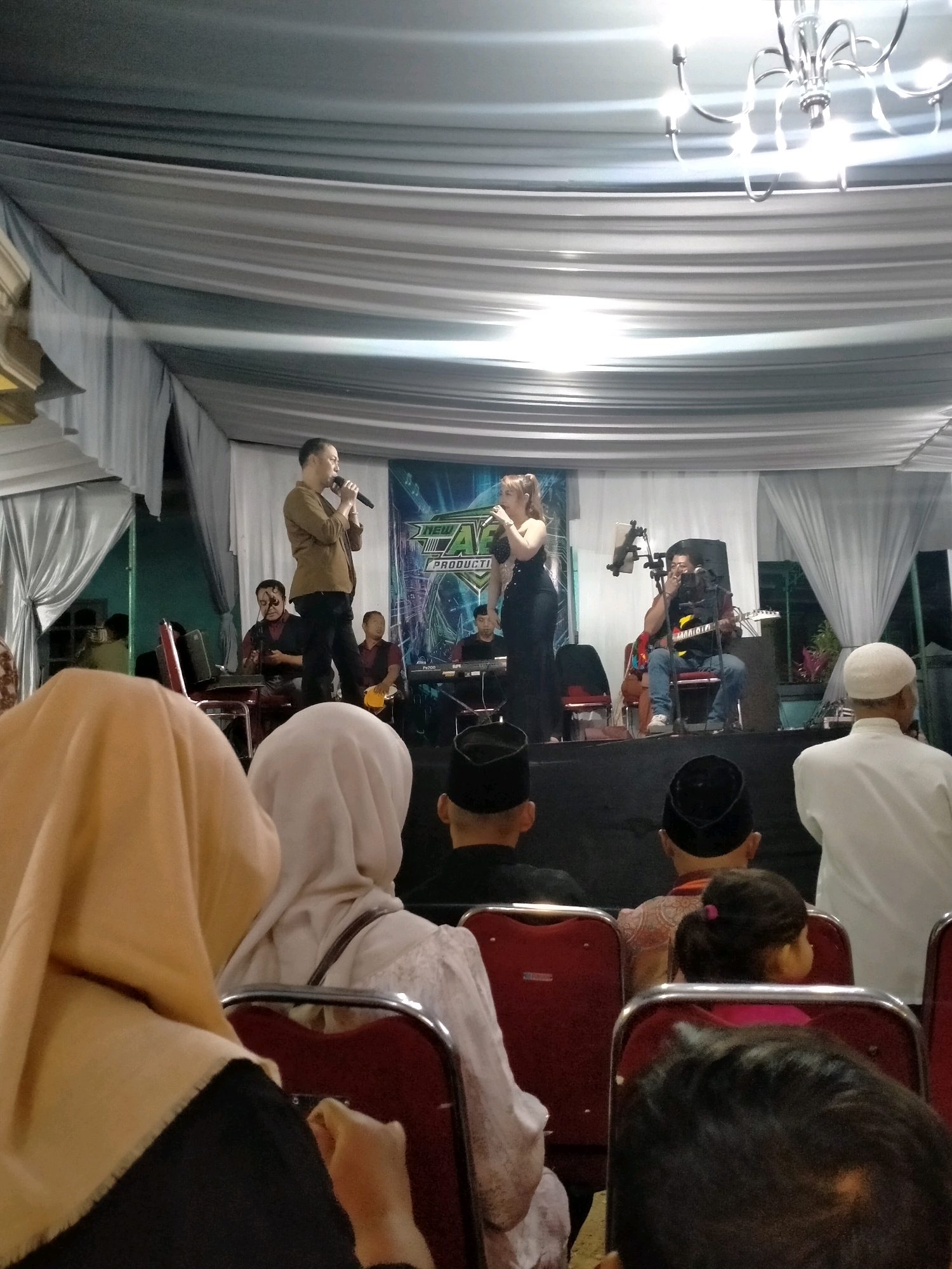 This image captures a live musical performance taking place under a draped white tent, likely at an outdoor event in Batu, Indonesia. The stage features a male singer in a brown shirt and black pants holding a microphone, and a female singer in a black outfit. Several band members are visible behind them, including a keyboardist and a guitarist. The audience is seated in rows of red chairs, with many wearing headscarves and traditional attire, indicating a cultural event. The foreground shows the backs of several audience members, including a child with their hair in a bun. The background is dominated by white drapes and a chandelier, creating a festive atmosphere. The overall scene suggests a lively and communal gathering, possibly a wedding or a local festival, held in an open area in Batu.
