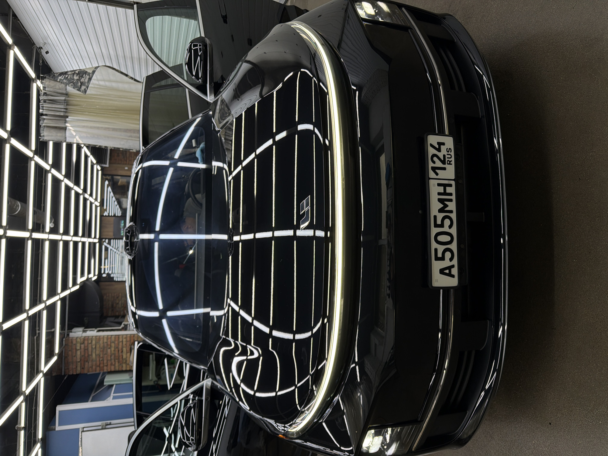 A close-up, rotated, and high-angle shot depicts the front of a dark-colored car in what appears to be a car detailing or washing facility. The car's license plate, "A505MH 124 RUS," is clearly visible, indicating the car is registered in Russia. The car's make is unknown, though its emblem is visible. The environment consists of a space with overhead grid lighting, common in such facilities to highlight imperfections in the car's paint. The reflection of the lights is visible on the car's hood, which is also shiny. Further, elements of the car wash interior can be seen in the car’s reflection. The location is likely in Krasnoyarsk, Russia, given the license plate code.