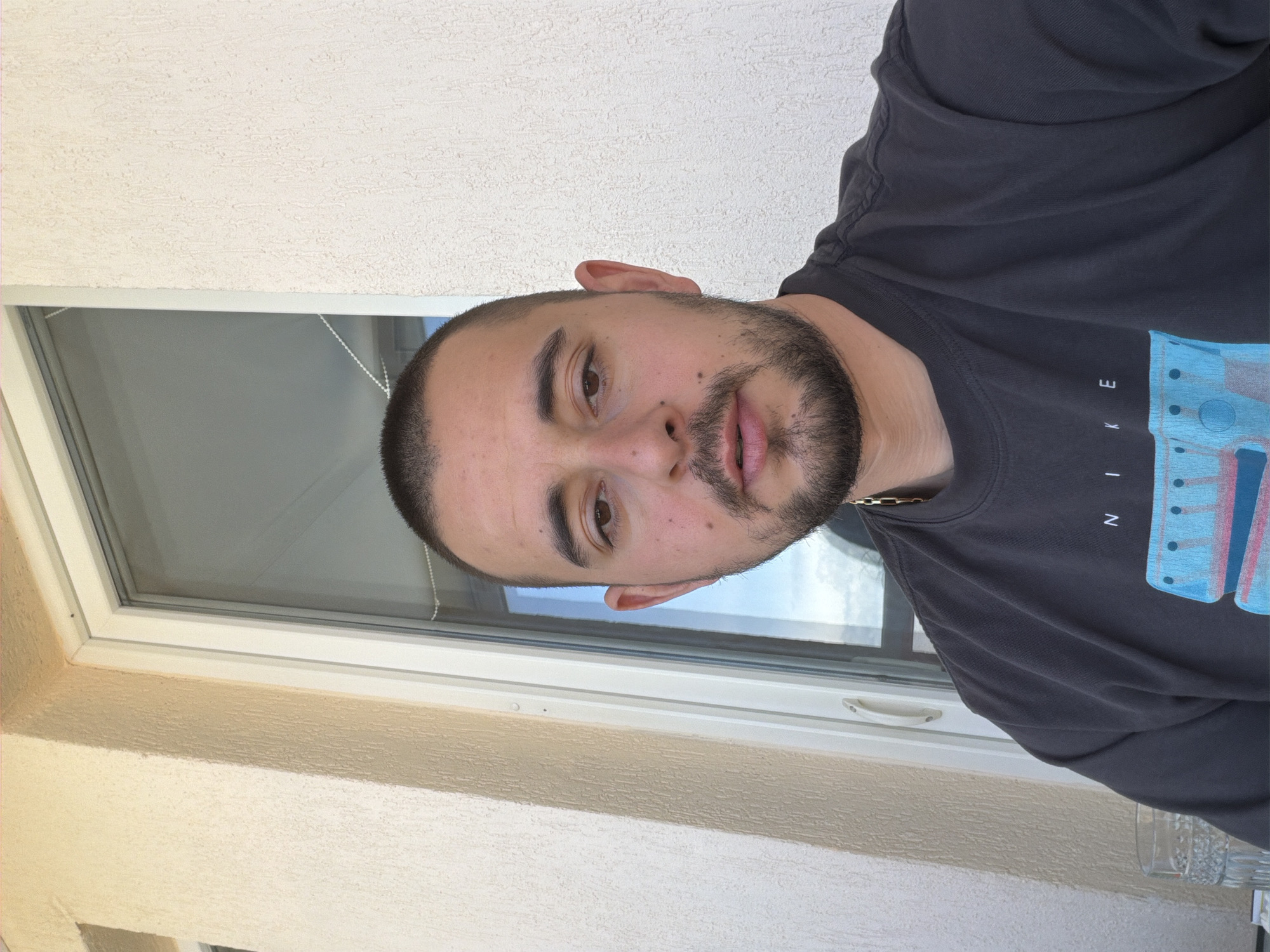 A man is shown in a close-up, waist-up view, positioned near a window with a white frame. He has a shaved head, a full beard, and is wearing a dark-colored t-shirt with light blue and pink graphic elements and the word "NIKE" printed on it. The wall behind him is light beige. The location is Unknown, Unknown.