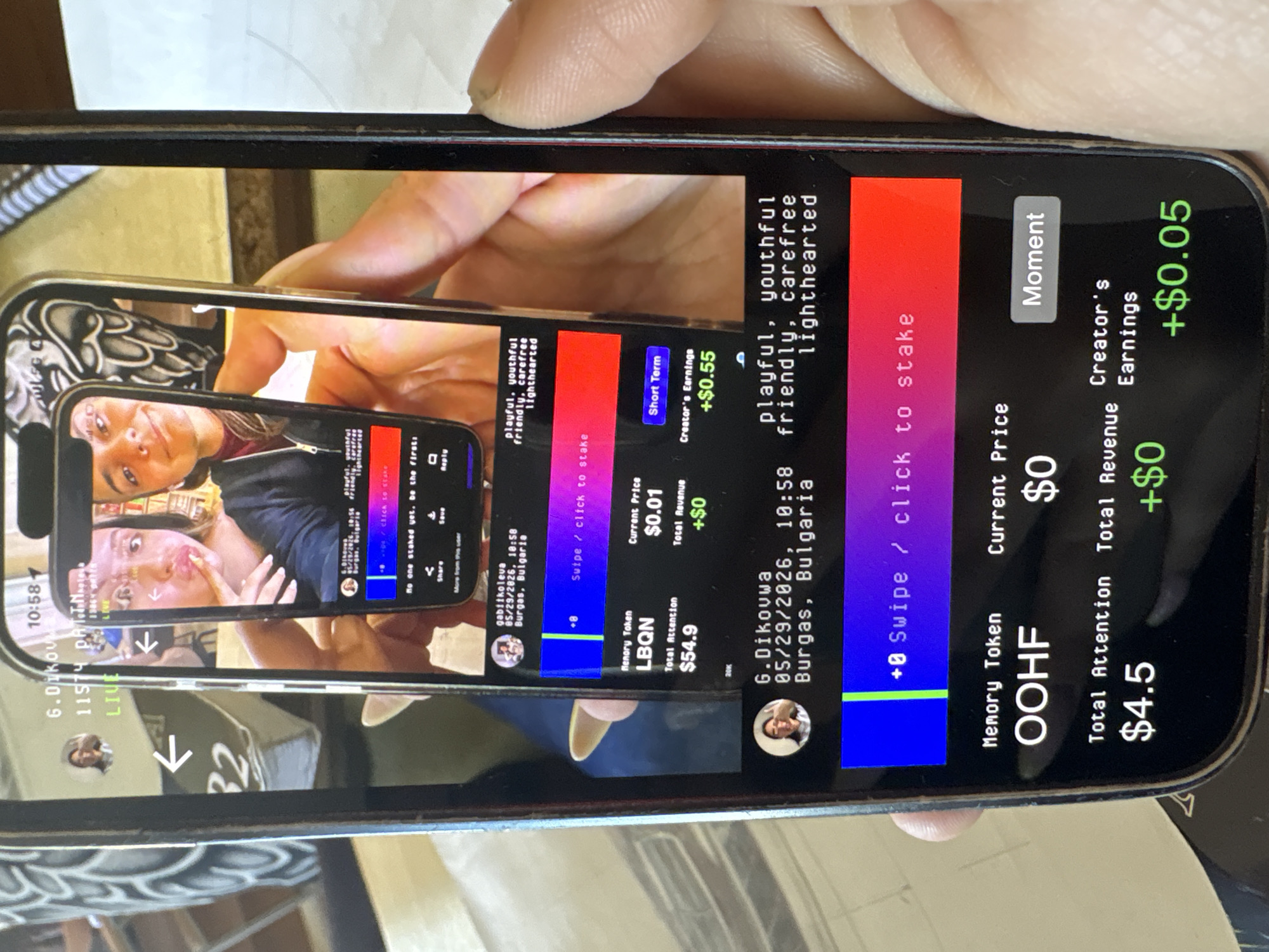 A hand holds a smartphone displaying a mobile app interface, mirrored repeatedly within the screen due to the phone's camera reflecting its own display. The app appears to be a platform for sharing and interacting with user-generated content, potentially involving cryptocurrency or digital tokens. The displayed content includes user profiles, dates (e.g., 05/29/2026, 10:58), location information (Burgas, Bulgaria), and various numerical values associated with tokens and earnings. The environment appears to be indoors, possibly on a wooden table or surface. The text on the screen also features descriptions of users or content as playful, youthful, carefree, and friendly. The tokens listed are LBQN and OOHF. The attention level for the OOHF token is $4.5 and its creator's earnings are $0.05.