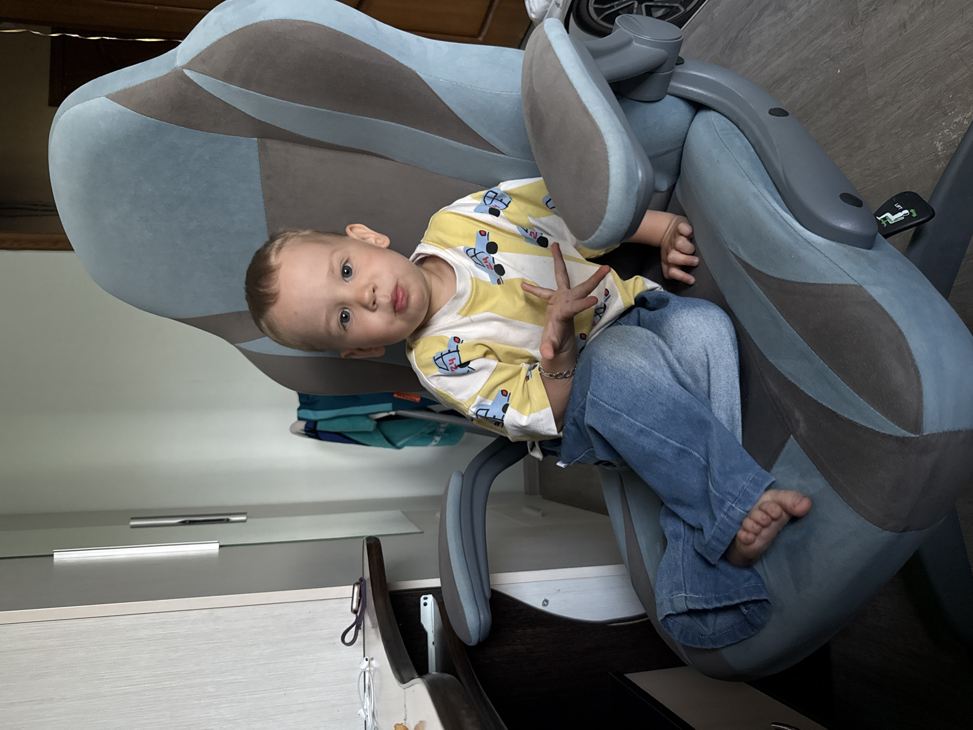 A young child is sitting in a blue and grey gaming chair. The child is wearing a yellow and white patterned shirt with blue jeans. The chair is positioned in a room with a white cabinet and a wooden door visible in the background. The floor is a dark wood laminate. The location is Rusanovo, Russia.
