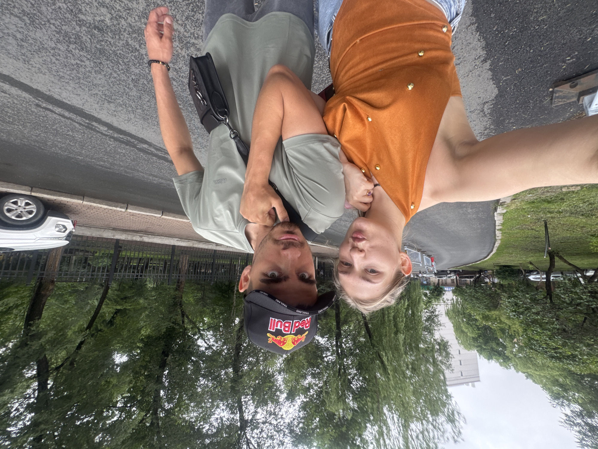 In this image, a young Caucasian couple is posing for a selfie outdoors. The woman, on the left, has blonde hair pulled back and is wearing an orange halter top with gold embellishments and blue jeans. She is puckering her lips as she holds the camera. The man, on the right, has dark hair and is wearing a green t-shirt and a black baseball cap with the Red Bull logo. He has a black crossbody bag slung over his shoulder. They are standing on a paved pathway next to a road. In the background, there are trees, a tall white building, and parked cars. A white SUV is visible on the right side of the frame. The scene appears to be in a park or residential area. The location is identified as Xiangfang District, China.