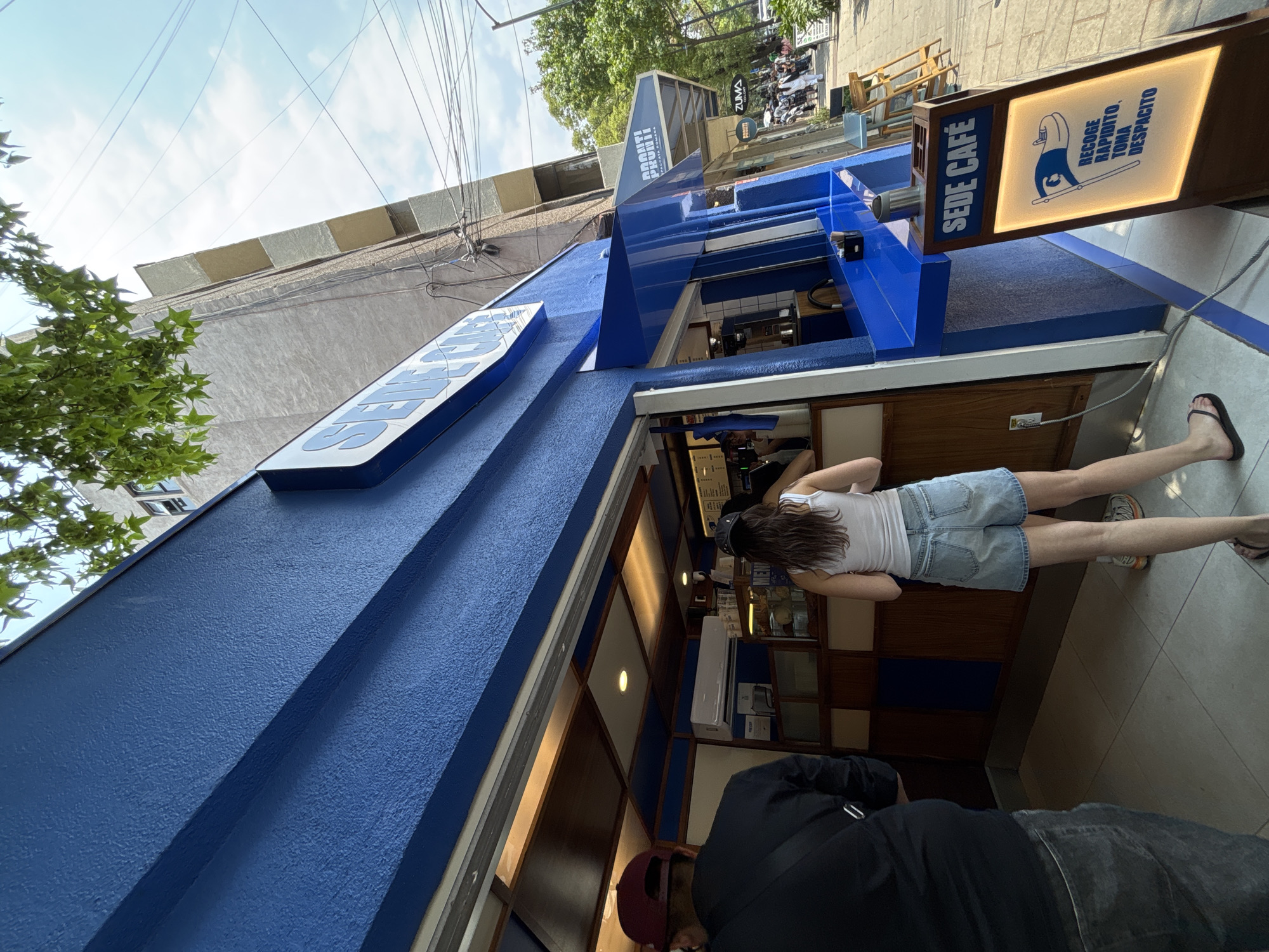 The image captures a scene at a cafe in Mexico City, Mexico. The cafe, named "Sede Café", is painted in a vibrant blue. Patrons are visible both inside and outside the establishment. A woman with brown hair, wearing a white tank top and denim shorts, is standing inside, interacting with someone behind the counter. Outside, a person dressed in a black shirt and a red hat is near the entrance. The environment includes the city's architecture, a street scene, and green trees, giving context to the cafe's location within Mexico City. The cafe has a sign featuring the phrase "RECOGE RAPIDITO, TOMA DESPACITO" (Pick up quickly, take it slowly).