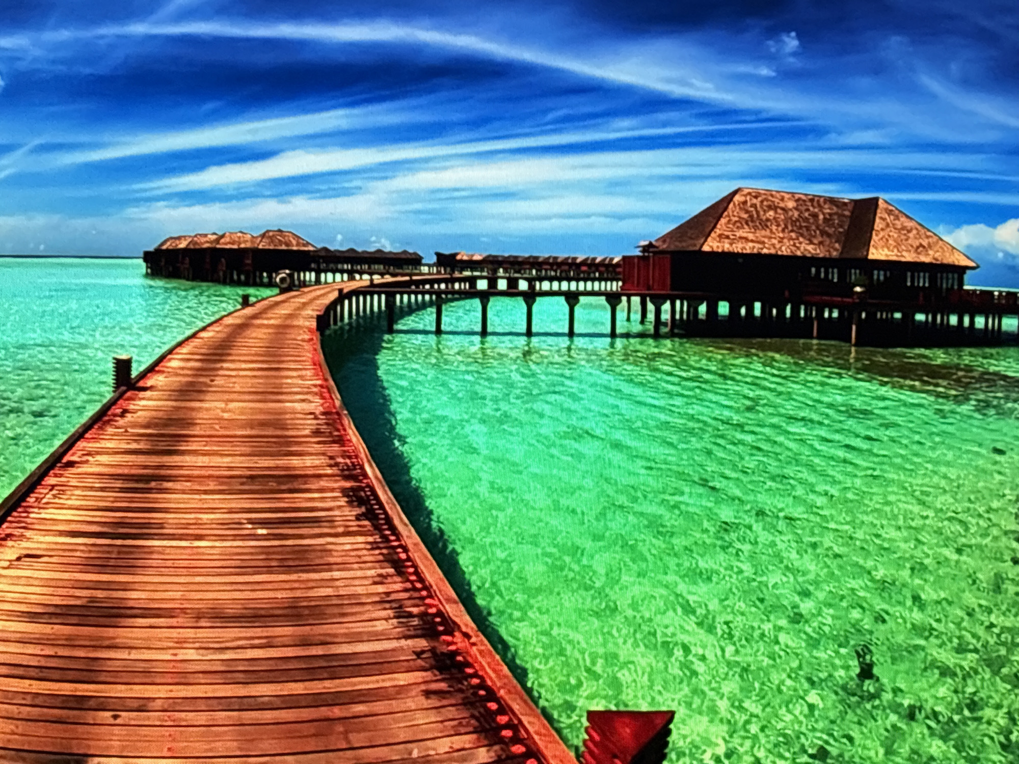 The image depicts an overwater bungalow resort situated in a tropical location, characterized by clear turquoise waters and a vibrant blue sky with wispy clouds. A wooden boardwalk curves towards the bungalows, which are built on stilts over the water. The bungalows themselves are dark wood with thatched roofs, and some are connected by similar boardwalks. The water is so clear that the supports of the structures are visible beneath the surface. The scene is serene and idyllic, evoking a sense of luxury and relaxation. The image is a photograph, likely taken during daylight hours. There are no people visible in the image. This location is not in Sofia, Bulgaria.