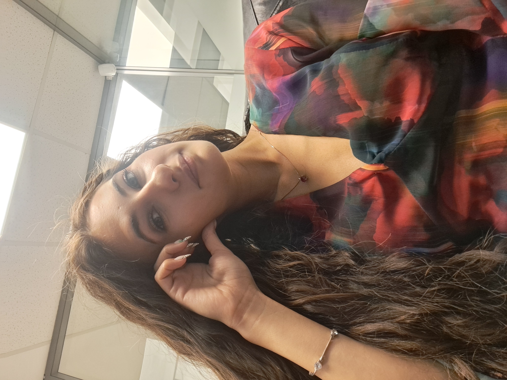 This image is a portrait of a young woman, likely a selfie, captured from a low angle looking upwards. The woman has long, wavy brown hair that extends across the frame. She has dark eyes and a calm, subtle expression, looking directly towards the camera. Her left hand is gently placed near her cheek, showcasing long, manicured nails with a distinctive white base and black dot pattern.
She is wearing a vibrant, multi-colored garment with an abstract pattern that incorporates shades of red, orange, green, blue, purple, and black, suggesting a light and flowing fabric. A delicate gold-colored necklace with a small, reddish-brown pendant adorns her neck, and a slender, silver-colored bracelet with small clear stones is visible on her right wrist.
The background indicates an indoor setting, possibly an office or a modern public space. A white security camera or sensor is mounted on a ceiling beam in the upper left corner. The ceiling features white acoustic tiles with a grid pattern. Bright natural light floods the scene from unseen windows or glass partitions, creating reflections in the background and illuminating the woman. A dark surface, possibly a chair or a partition, is visible behind her right shoulder. The overall lighting suggests it is daytime. There is no visible text in the image.
