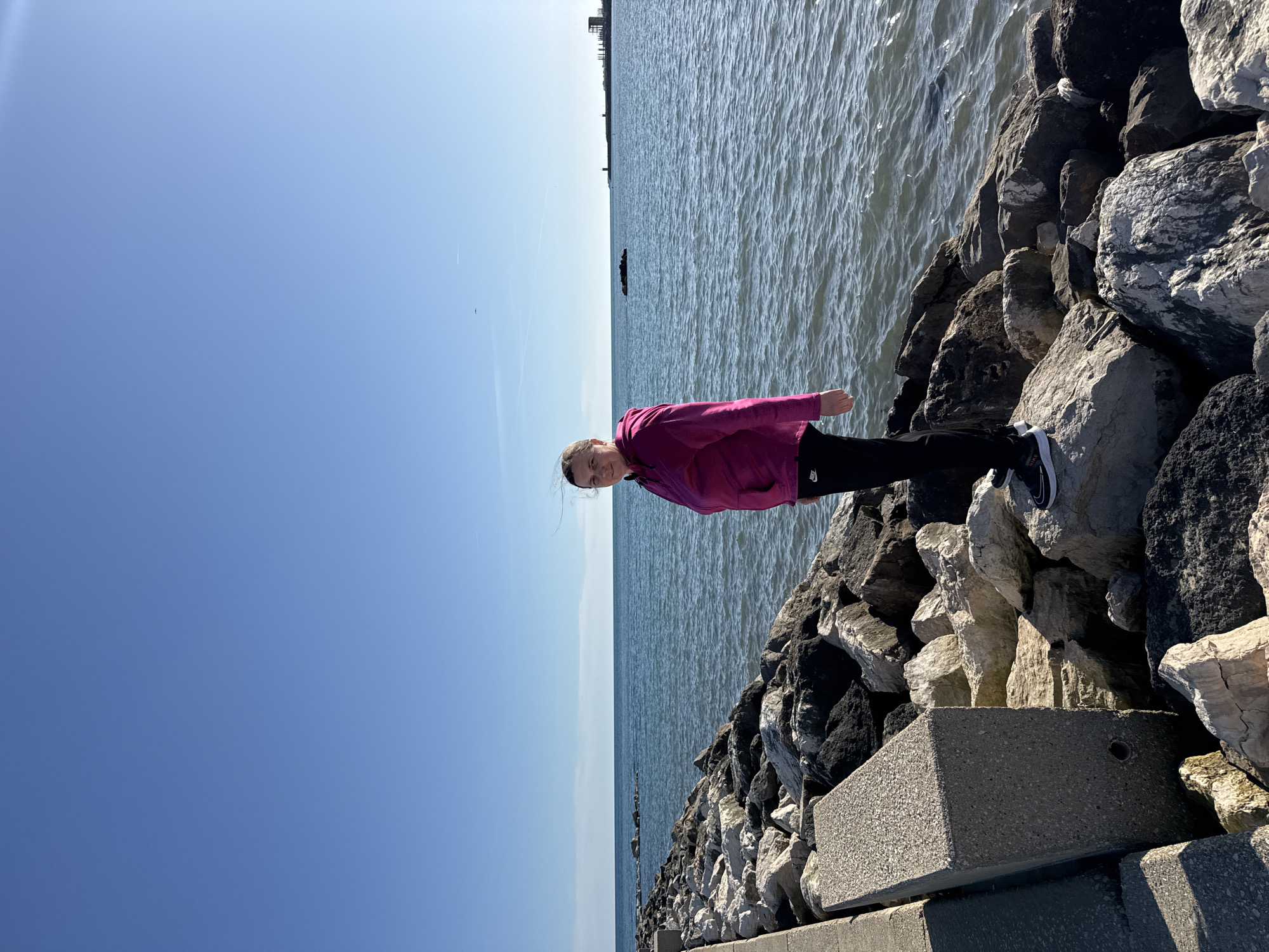 A young person, likely a girl, is captured standing on a rocky breakwater in Nettuno, Italy, under a vast, clear blue sky. She is positioned towards the left of the frame, wearing a bright magenta or pink jacket and dark pants, with a small white logo (possibly Nike) visible on her left pant leg. She faces the camera with a calm expression.
To her right, the blue waters of the Mediterranean Sea stretch to the horizon, showing gentle ripples and reflecting the bright sunlight. The breakwater, composed of numerous grey and white boulders, forms a rugged line extending into the distance along the right side of the image, separating the land from the sea. In the immediate foreground, a grey concrete block is visible on the breakwater to the person's left.
The bright, even lighting and the cloudless sky indicate a sunny day, likely mid-day. Faint, distant structures are visible on the horizon line across the water. High in the sky, a faint contrail is present, along with a tiny dark speck that could be a distant bird. The scene depicts a serene coastal environment with calm weather conditions.