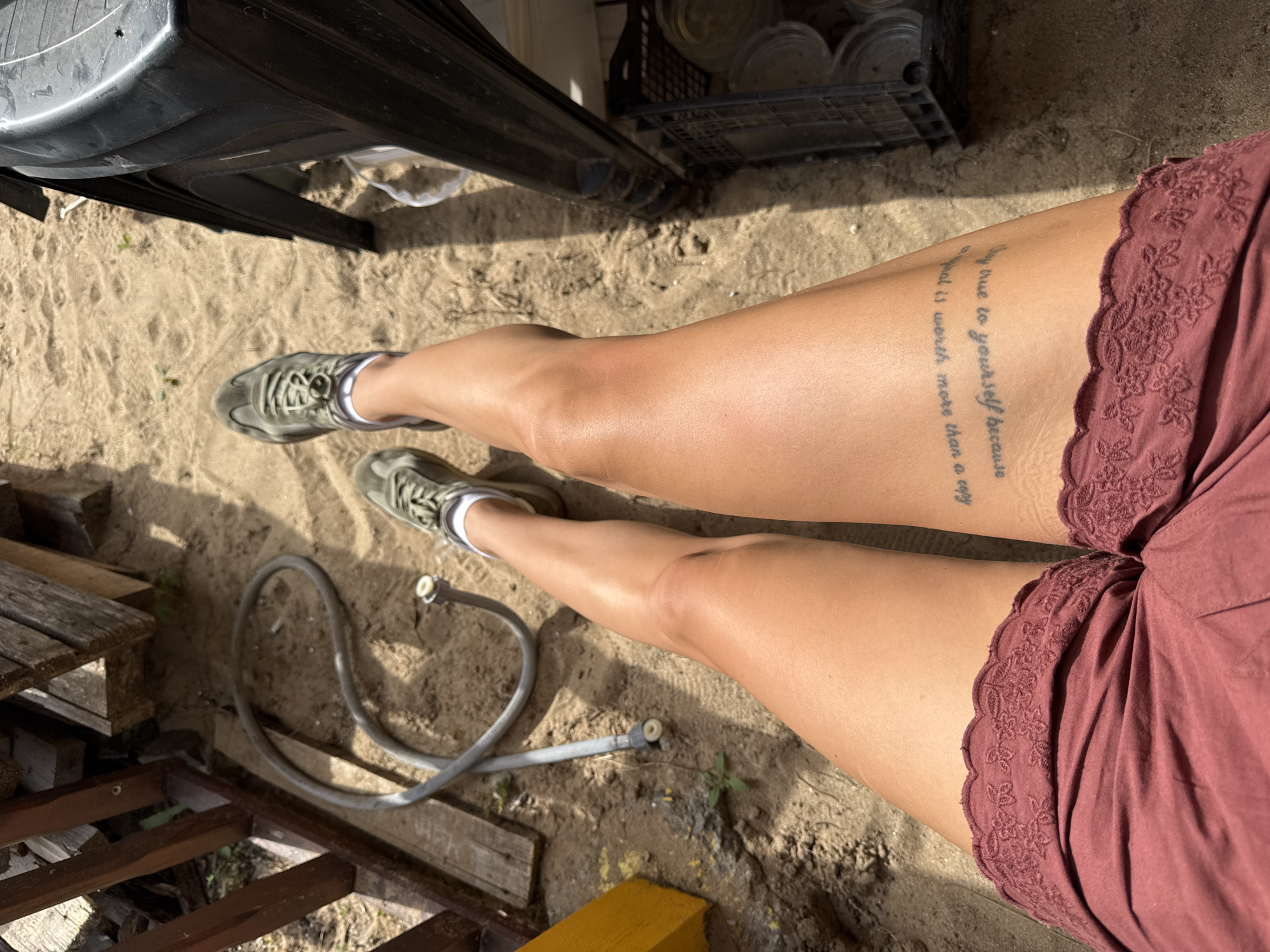 This media file features a close-up shot of a person's legs and thighs, seen from a high angle. The individual is wearing olive green sneakers and a rust-colored garment with lace trim. A tattoo with text is visible on the left thigh. The setting appears to be outdoors, on sandy ground, with wooden structures and a hose present. In the background, a black plastic crate filled with glass jars is partially visible. The lighting suggests it is daytime, possibly sunny. The overall impression is casual and relaxed. The text on the tattoo is partially readable and appears to be a quote: "stay true to yourself because a one in a million is worth more than a copy". The location is identified as Sozopol, Bulgaria.