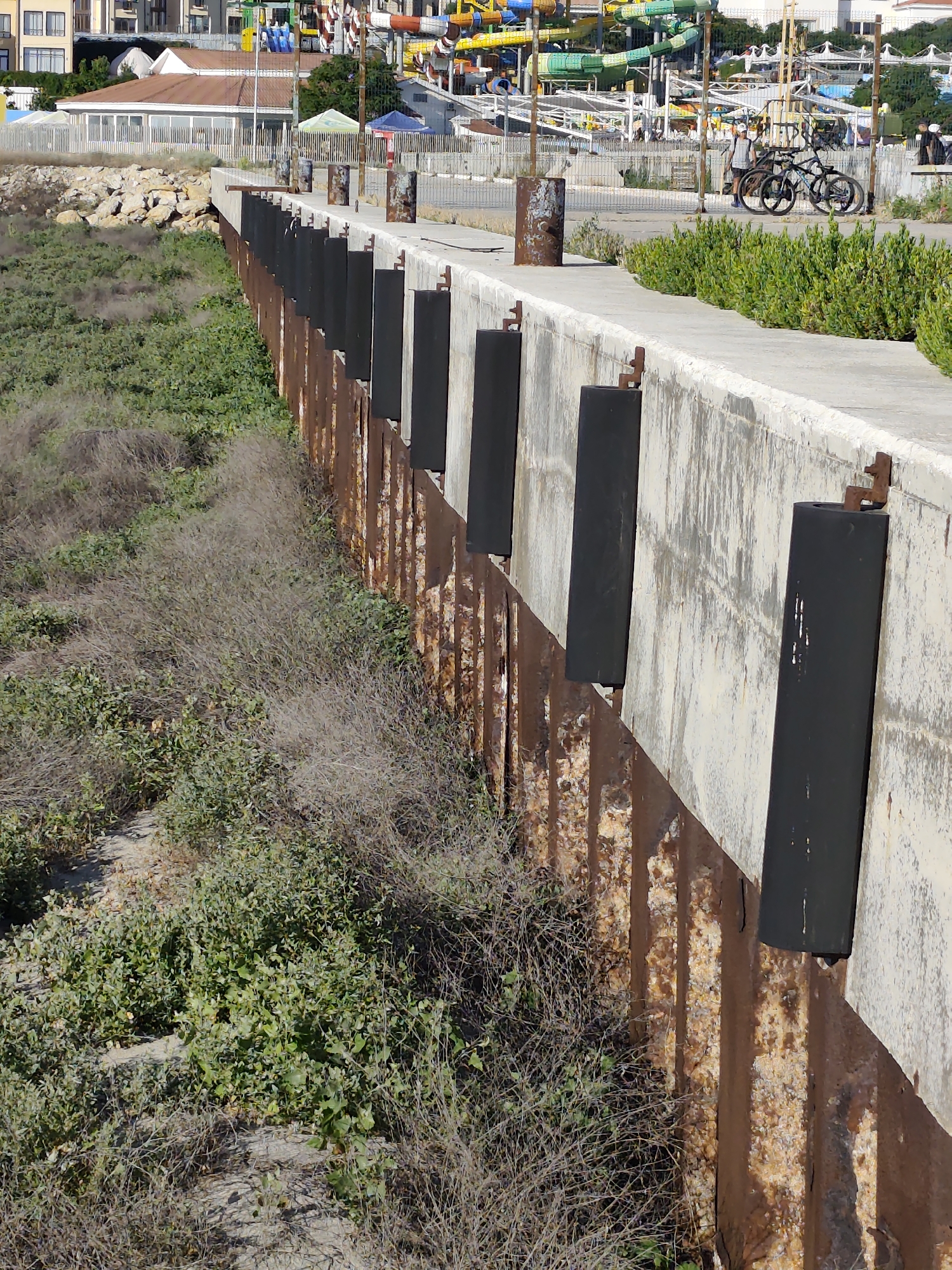 This media file depicts a section of a weathered concrete seawall, possibly in Aktau, Kazakhstan. The wall is reinforced with rusty corrugated metal sheets and features several black rectangular fenders mounted at intervals. The top of the wall is a concrete walkway. To the left of the wall, the ground slopes down, covered in a mix of dry, scrubby vegetation and patches of green, low-lying plants.
In the background, beyond the walkway, there's a glimpse of a building with light-colored walls and a white railing, suggesting a developed area adjacent to the waterfront. Further in the distance, a colorful water park with slides and structures is visible, hinting at a recreational or tourist destination. A few people can be seen near the water park and along the walkway. Two bicycles are parked on the walkway near some bushes.
The scene appears to be set during the daytime, with good lighting. The presence of the water park suggests a warm weather activity. The overall impression is one of a functional coastal structure bordering a leisure area, with a blend of natural and man-made elements. There is no visible text in the image.